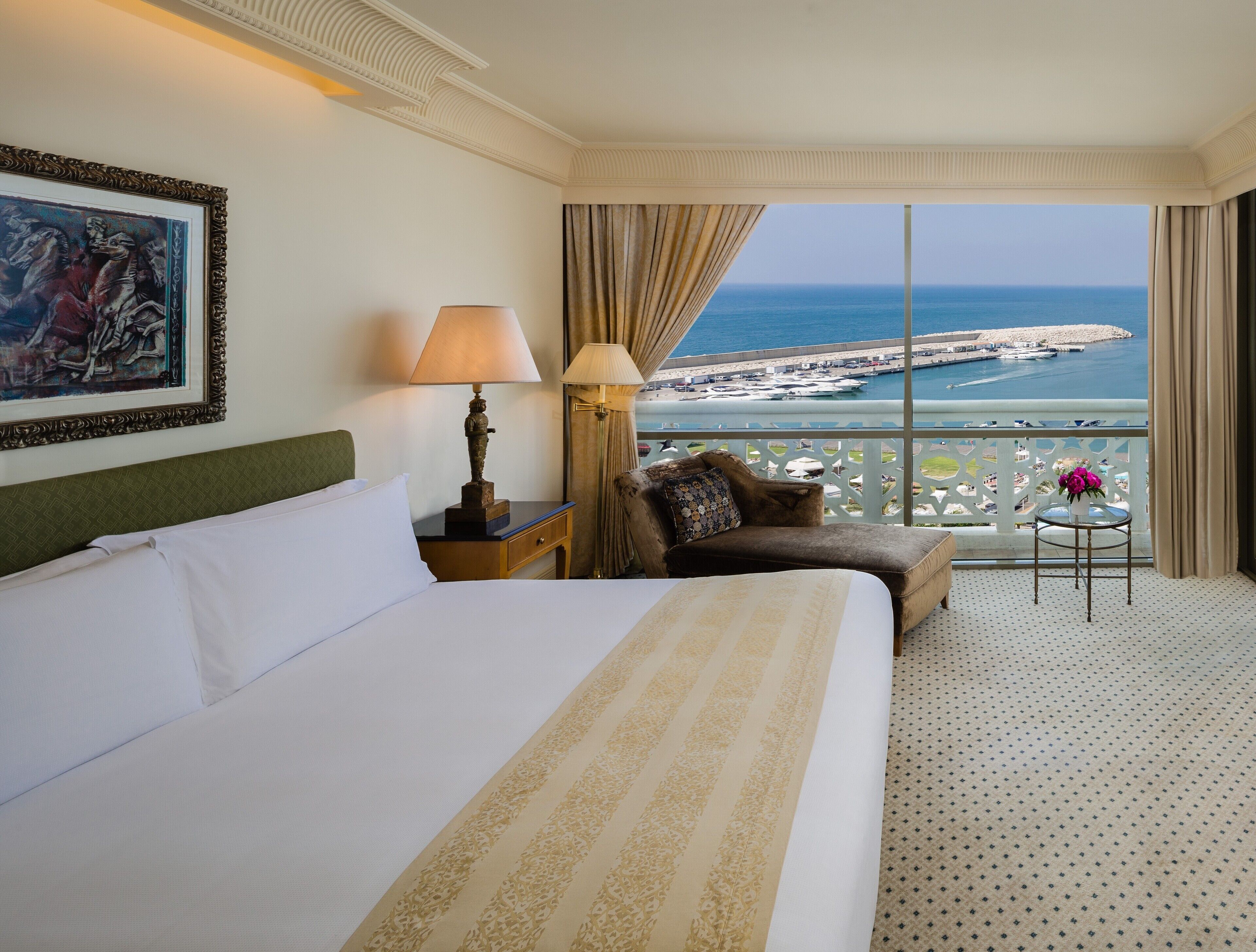 A hotel room with a large bed, a nightstand with a lamp, a sofa, a small table with a vase, and a view of the sea and a pier.