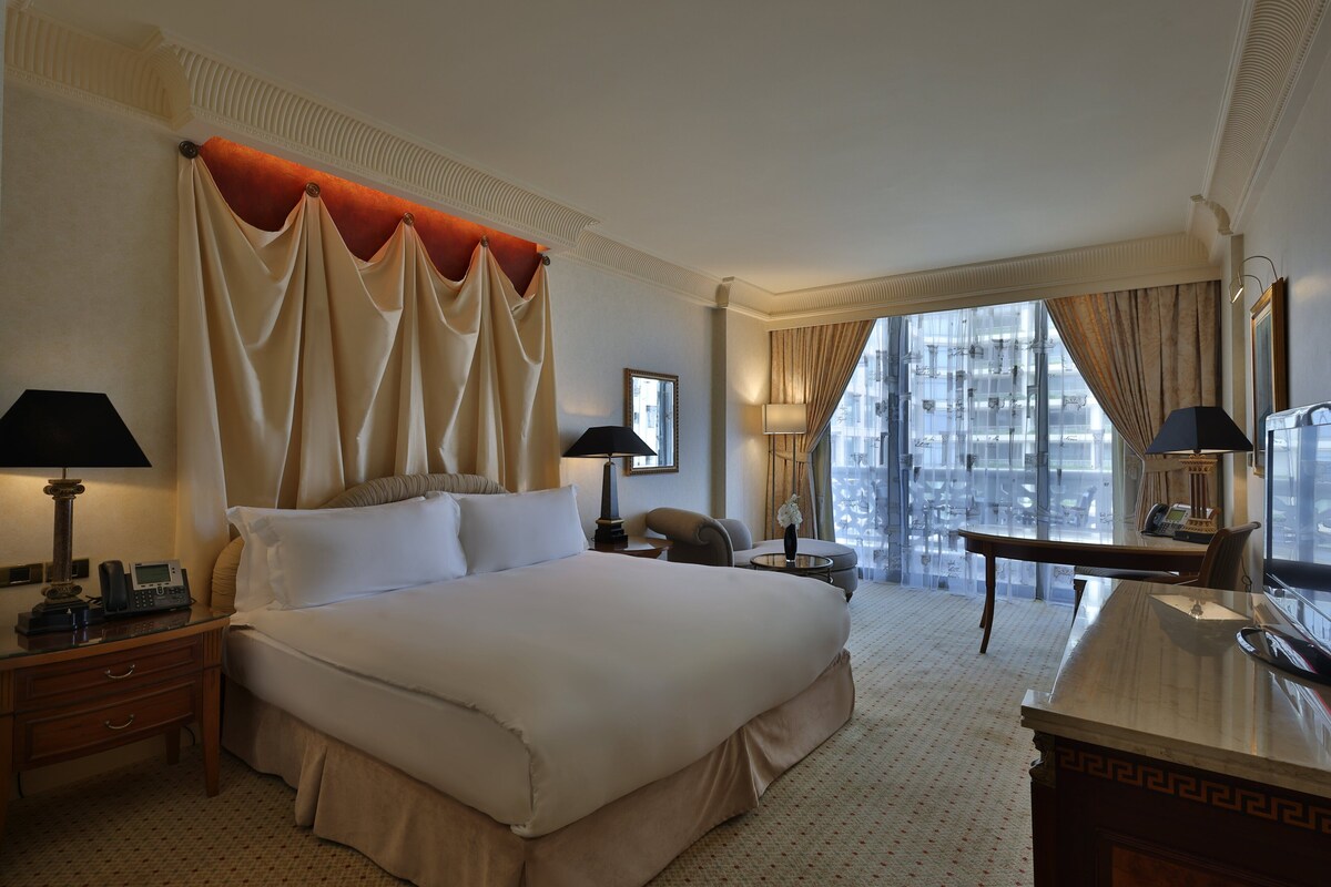 Habitación de hotel con una cama grande, un escritorio, una silla y vistas a la ciudad a través de amplios ventanales.