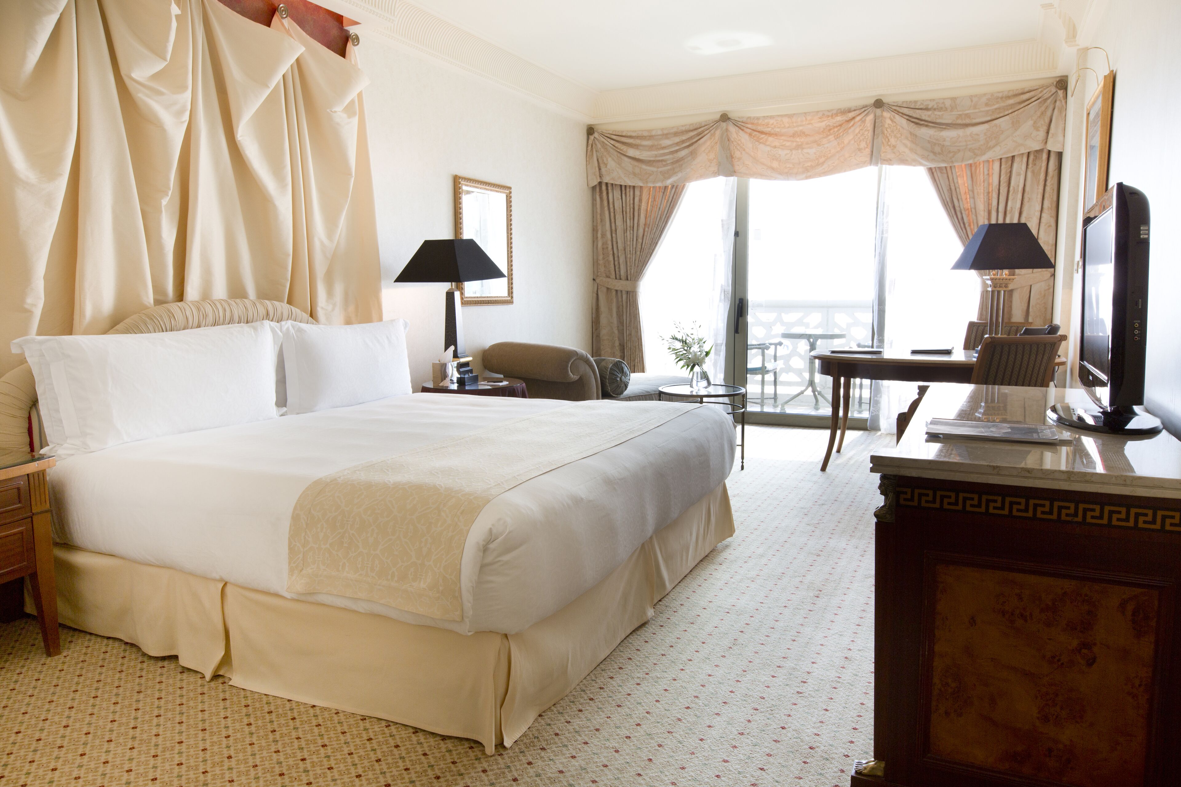 Habitación de hotel con una cama grande, un escritorio con silla, televisor y balcón con cortinas.