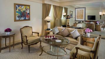 A hotel room with a sofa, armchairs, a glass coffee table, a desk with a lamp, and artwork on the walls.