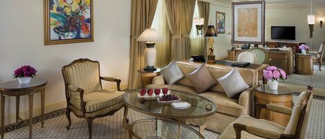 Habitación de hotel con sofá, sillones, una mesa de centro de vidrio, escritorio con lámpara y obras de arte en las paredes.