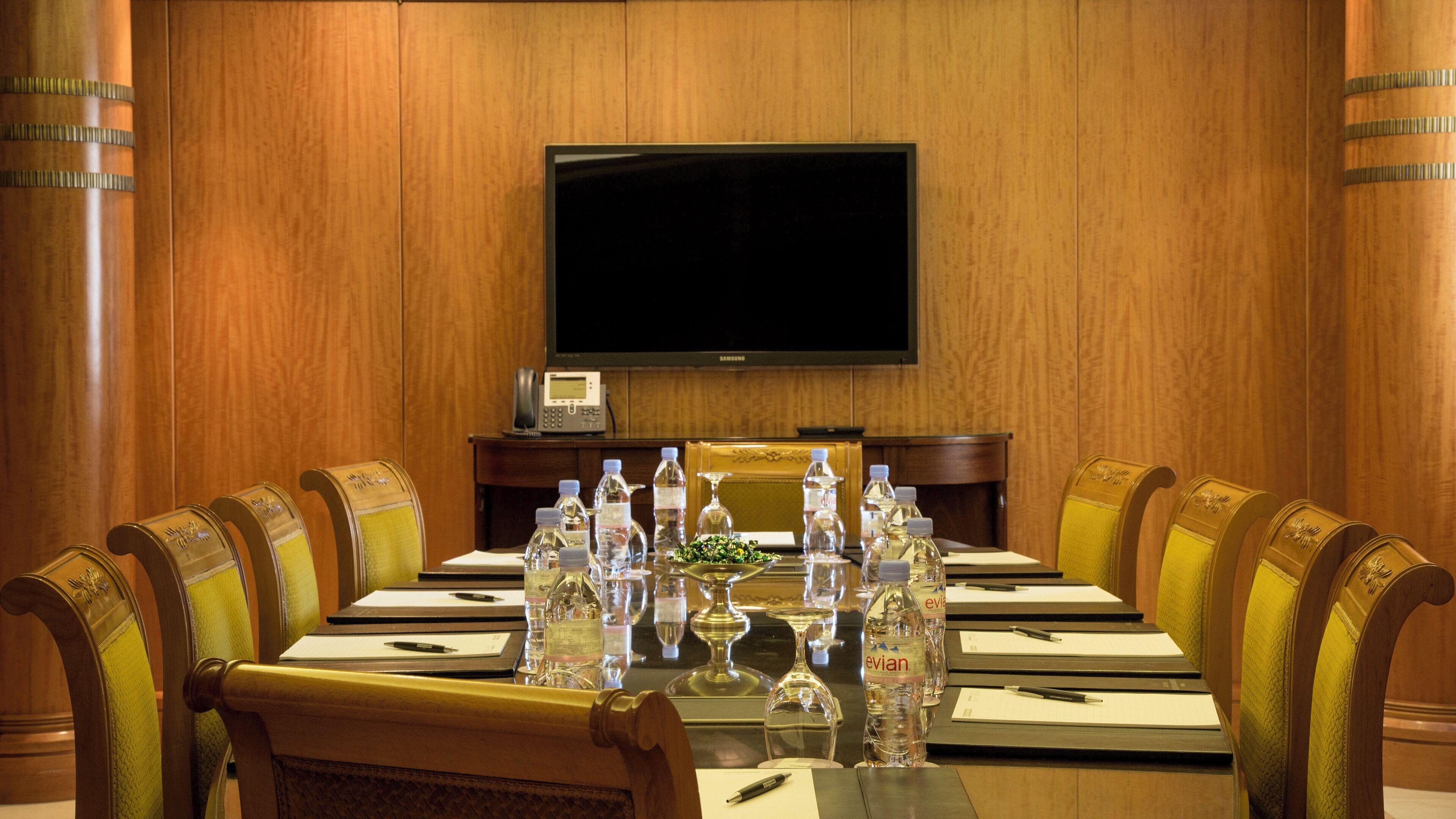 A conference room with a long table, chairs, a flat-screen TV, and a telephone on a wooden panel wall.