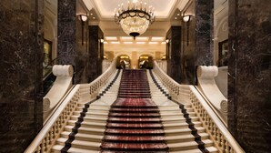 A grand staircase with marble walls and a chandelier, leading to a grand hall.