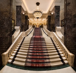 A grand staircase with marble walls and a chandelier, leading to a grand hall.
