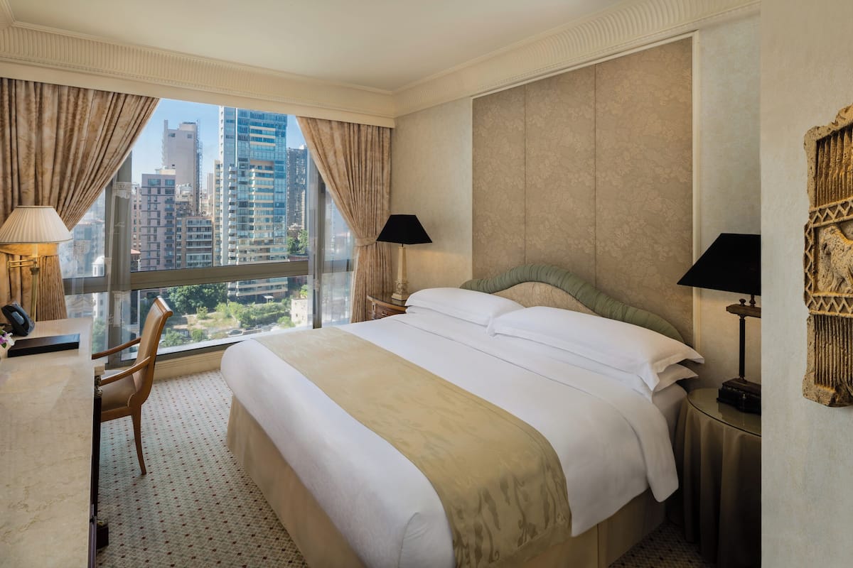Une chambre d’hôtel avec un grand lit, deux lampes de chevet, un bureau avec un téléphone, une chaise et une vue sur de hauts immeubles par la fenêtre.