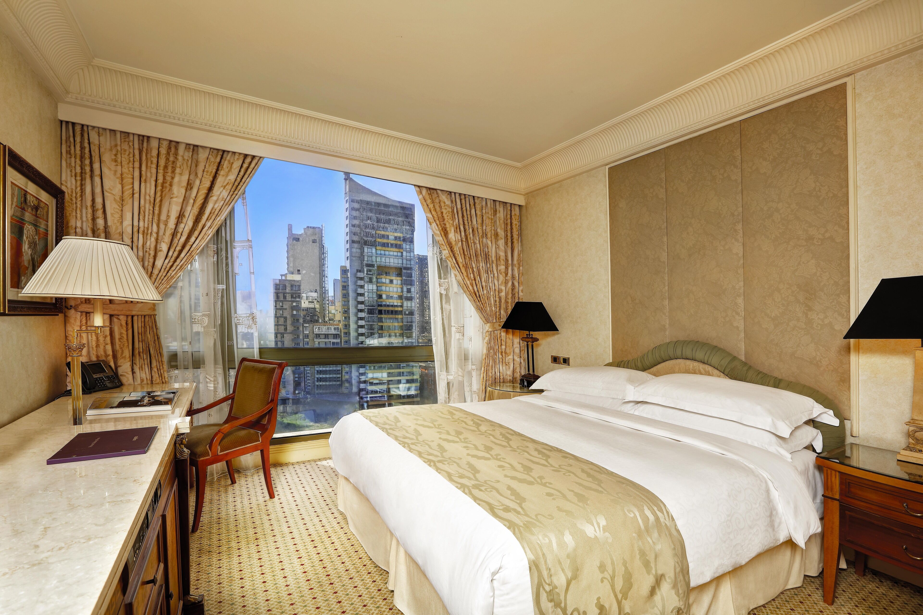 Quarto de hotel com uma cama grande, duas mesas de cabeceira, uma escrivaninha com cadeira e vista para a cidade através de amplas janelas.