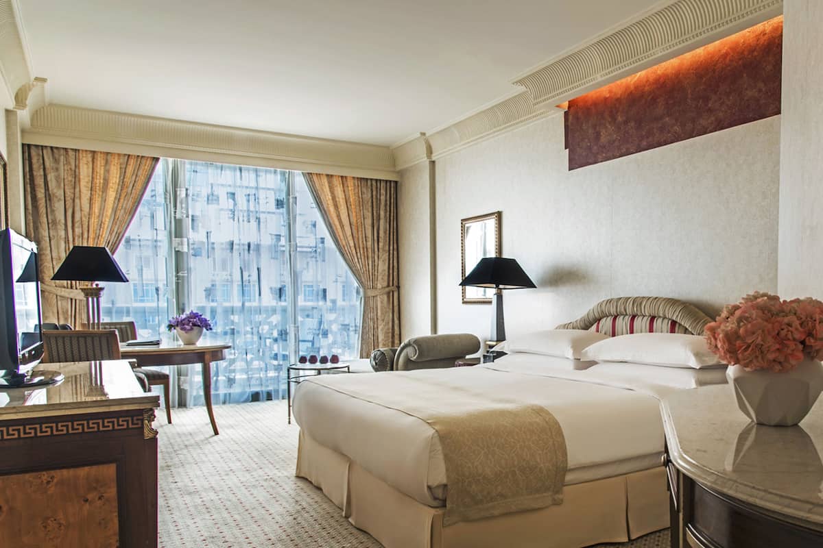 Une chambre d’hôtel avec un grand lit, un bureau avec une lampe, une chaise, un vase de fleurs et une vue sur les bâtiments par la fenêtre.