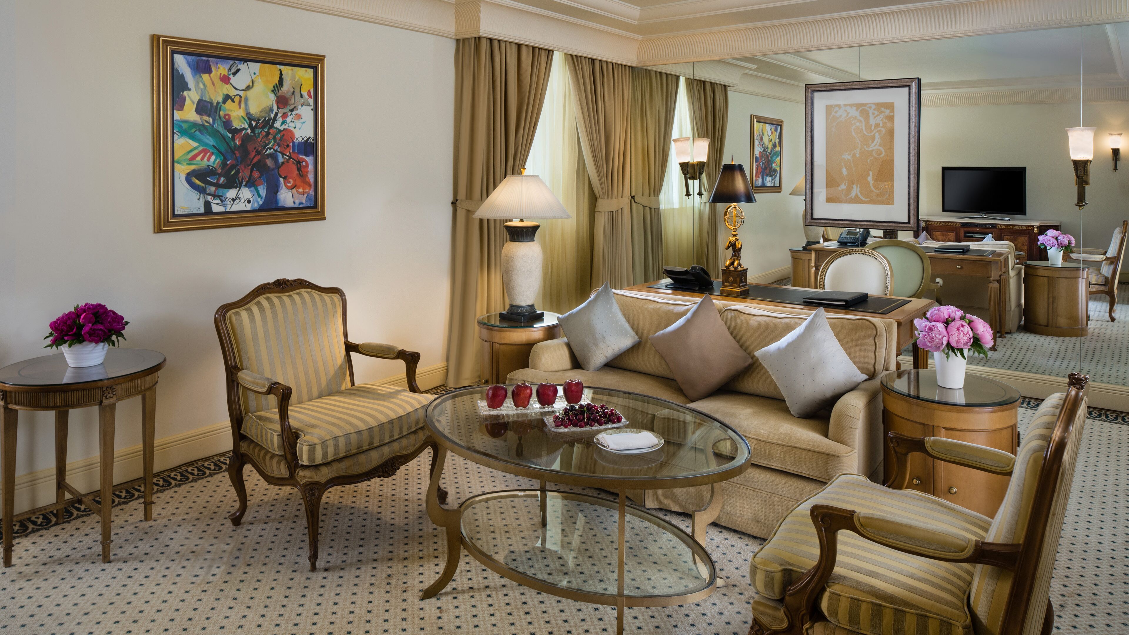 A well-appointed hotel room with a sofa, armchairs, a glass coffee table, and a desk area with a television and telephone.