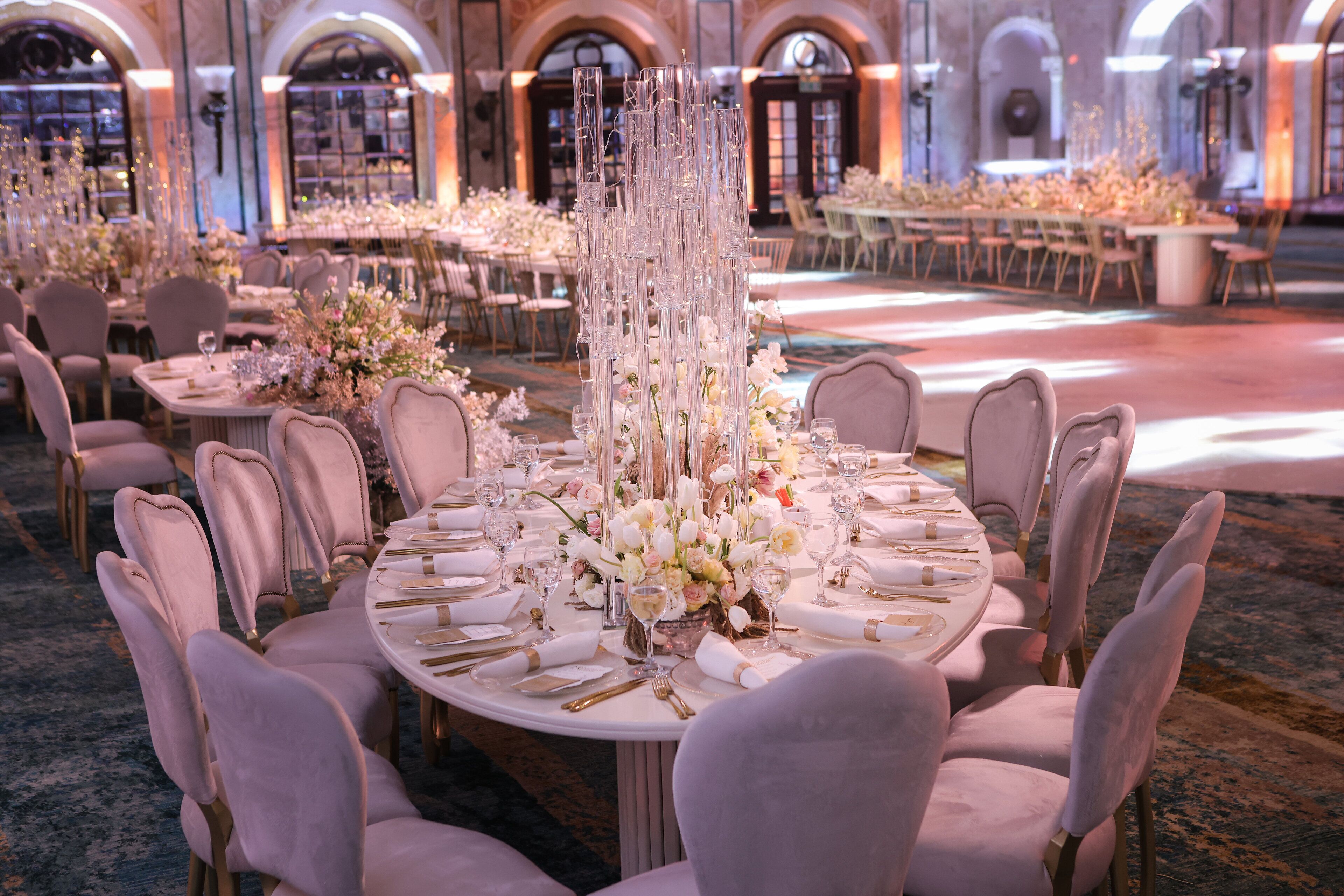 A banquet hall with round tables set for a formal event, featuring tall floral arrangements and elegant table settings.