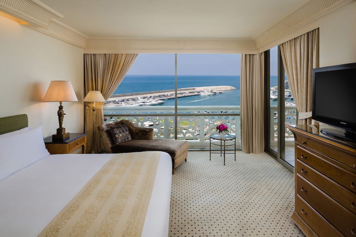 Une chambre d’hôtel avec un grand lit, un canapé, une télévision et un balcon offrant une vue sur la mer et les bateaux.