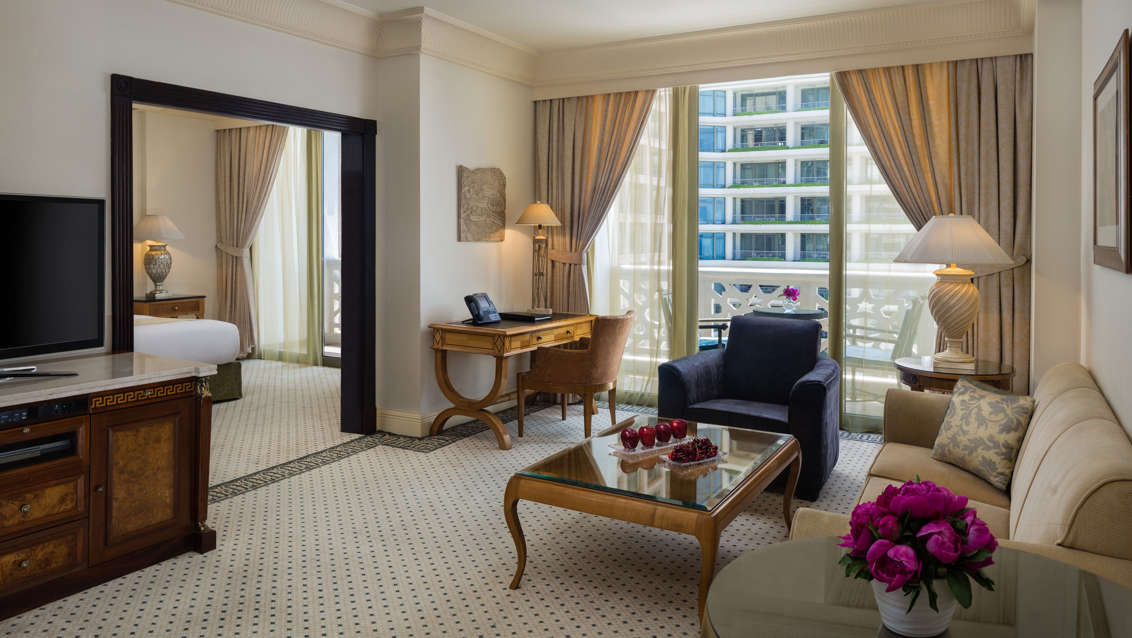 A hotel room with a large bed, a desk with a computer, a TV, a coffee table with flowers, and a view of the city.