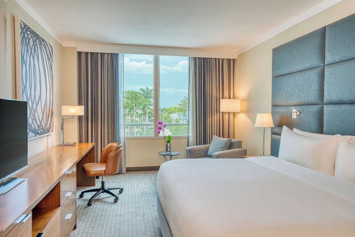 A hotel room with a bed, two chairs, a desk, a TV, and a window with curtains.