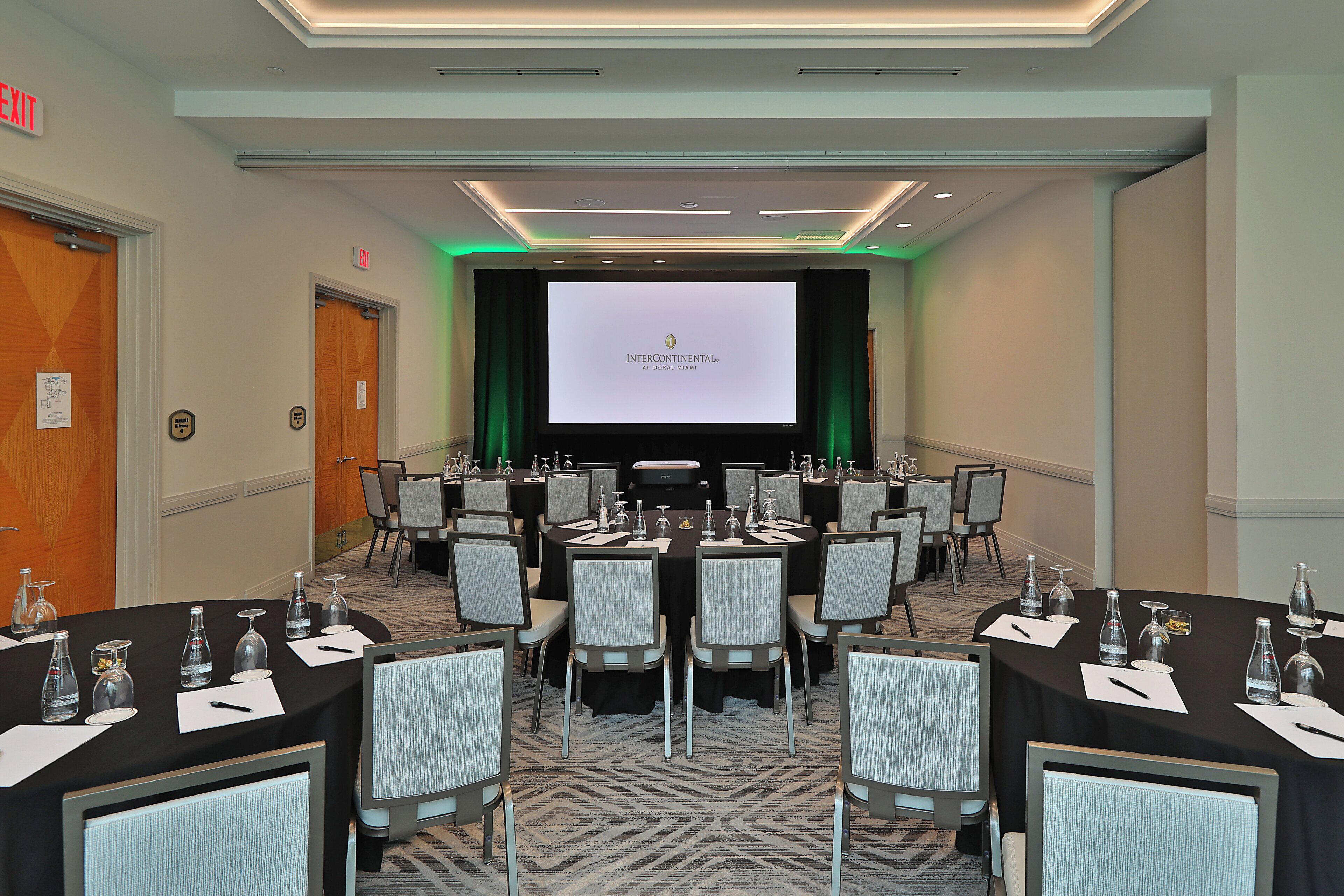 Un salón de conferencias con varias mesas, sillas y una pantalla grande que muestra 'INTERCONTINENTAL AT DORAL MIAMI'.