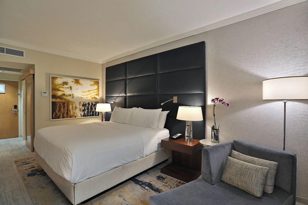 Een hotelkamer met een groot bed, twee bedlampen, een klein tafeltje met een telefoon, een grijze fauteuil en een schilderij aan de muur.
