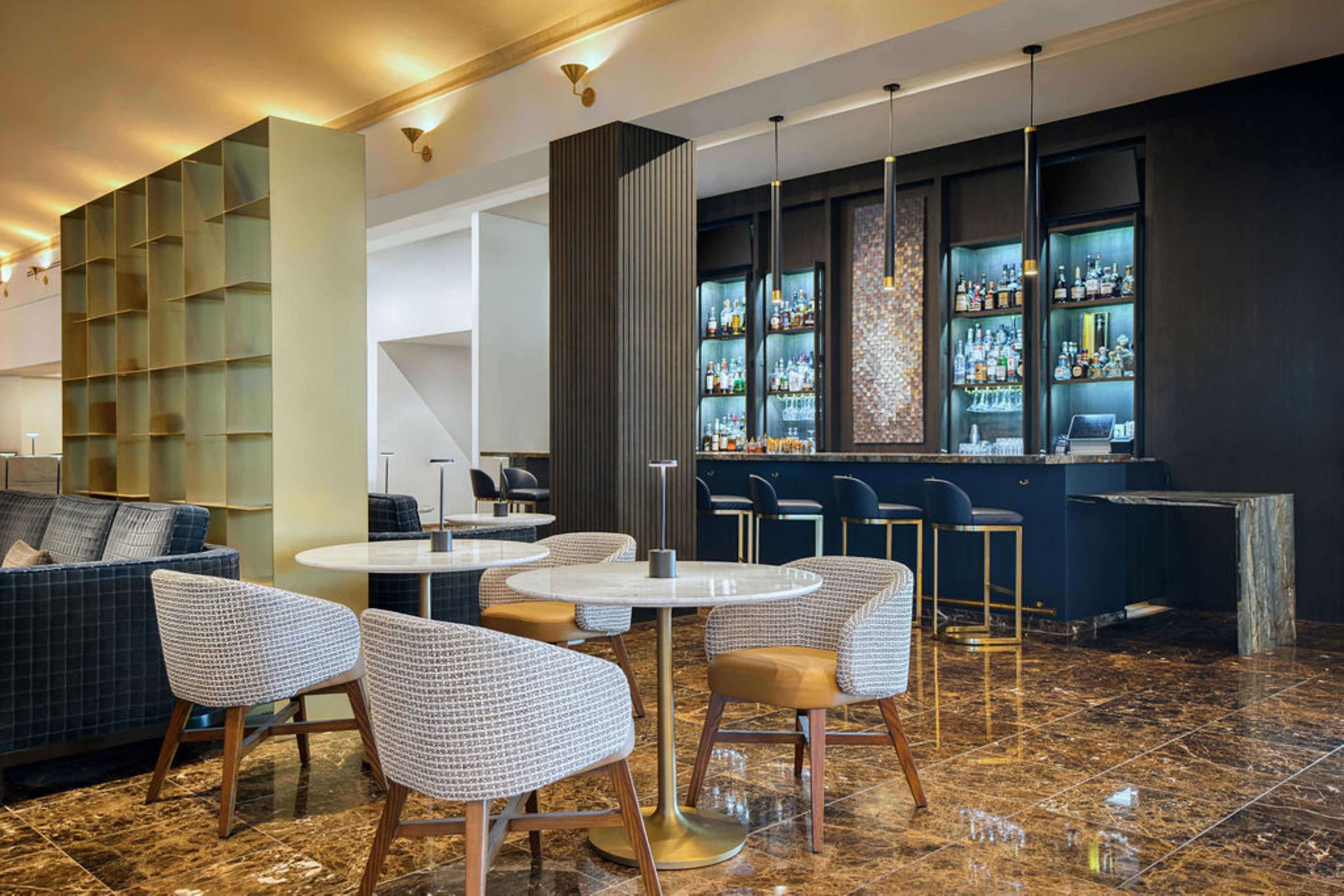 Zona de bar con mesas redondas, taburetes y una barra con estantes llenos de botellas.