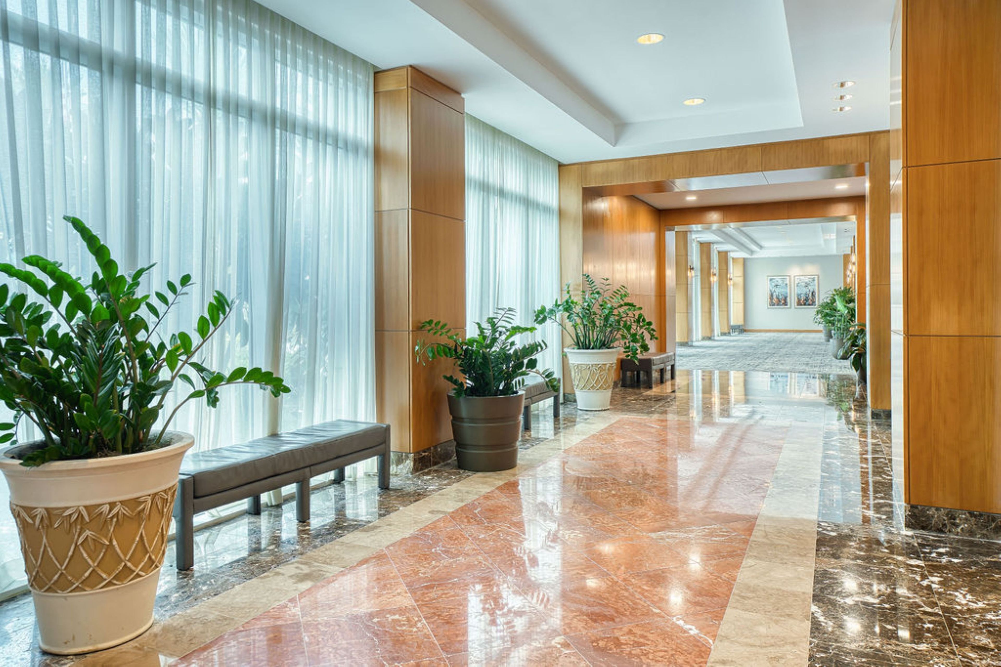A hotel corridor with marble flooring, potted plants, a bench, and a view of the lobby area.