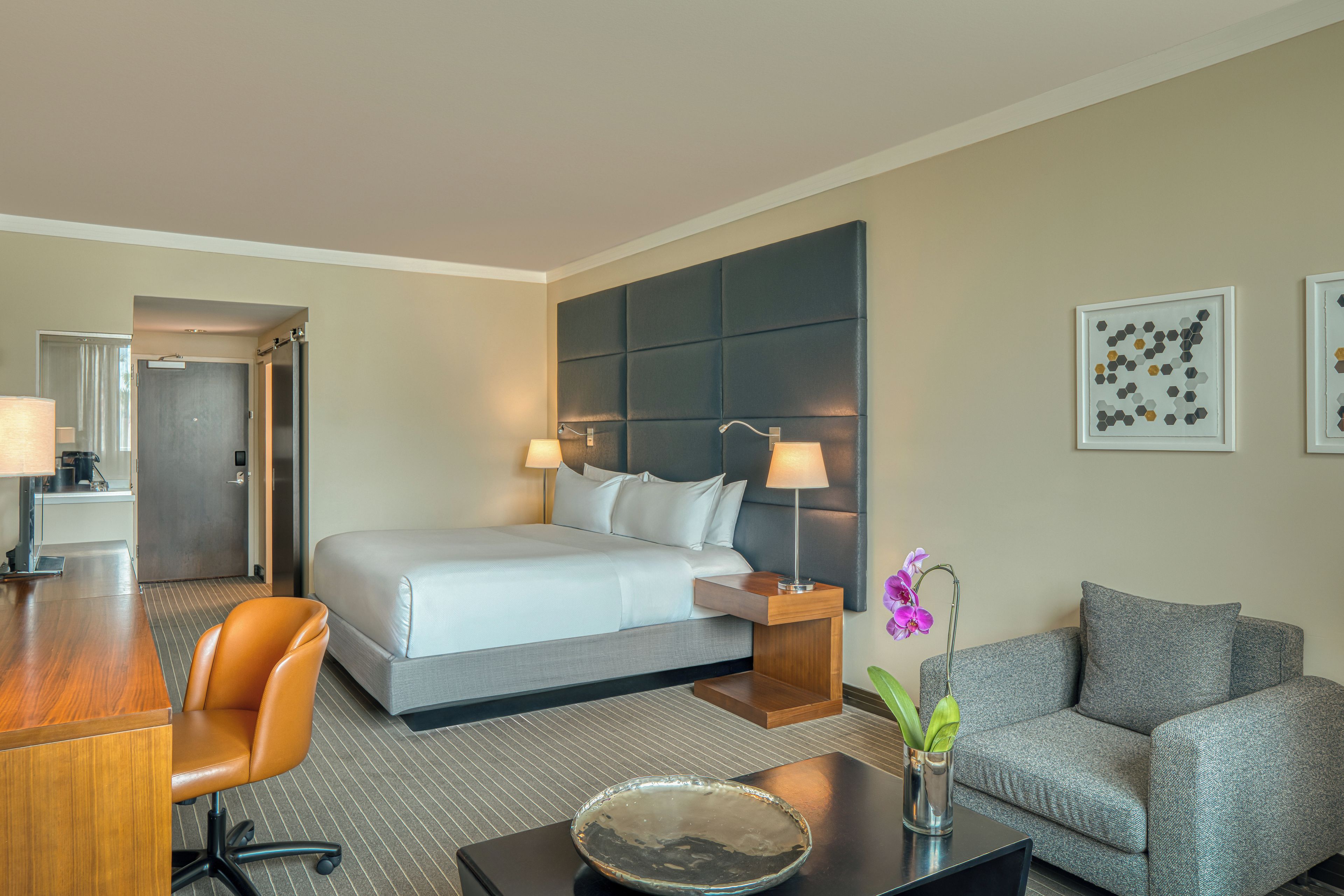 A hotel room with a bed, bedside tables, a desk, a chair, a sofa, and two framed artworks on the wall.