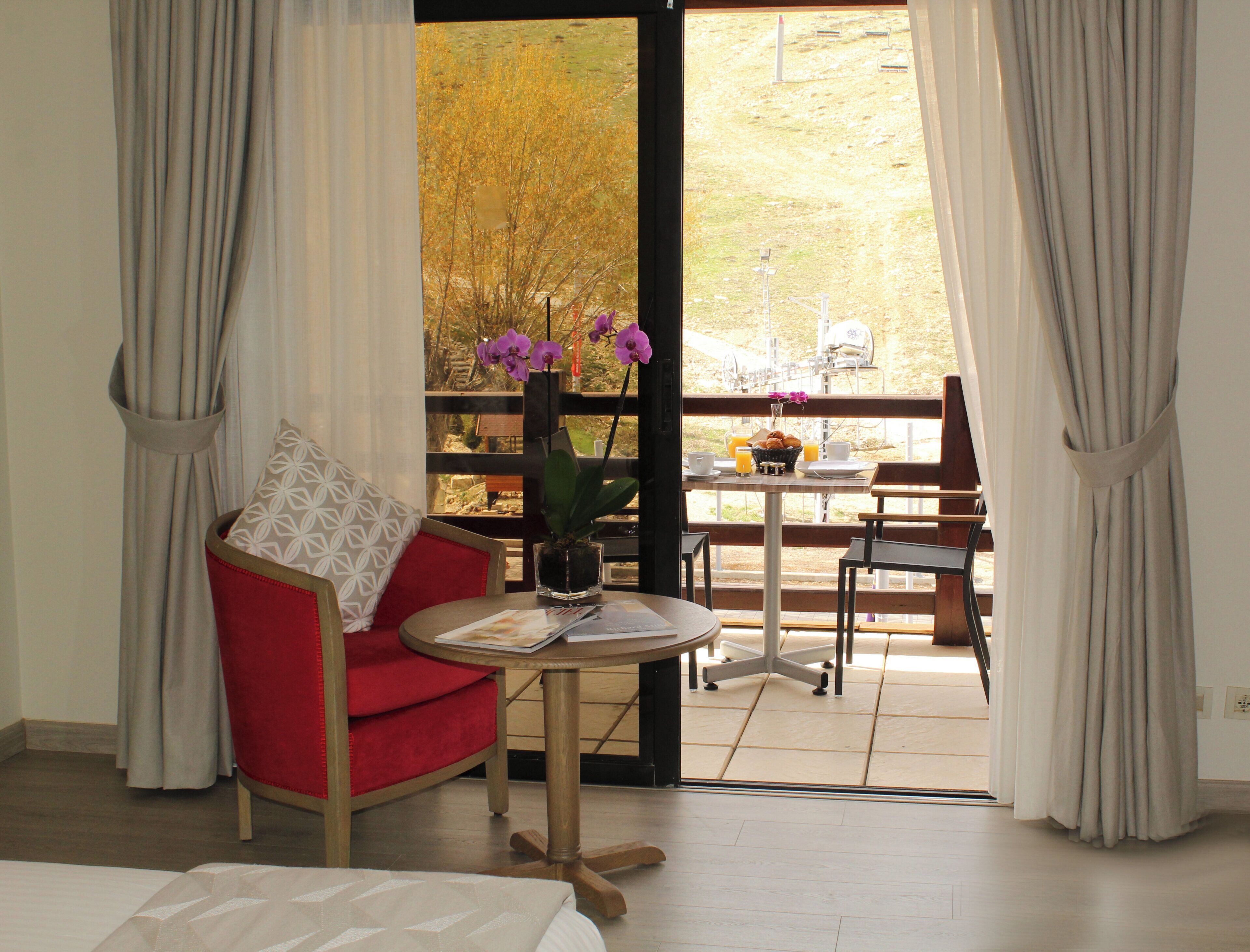 A room with a red chair, a small table with a vase of flowers, and a view of the outdoors.