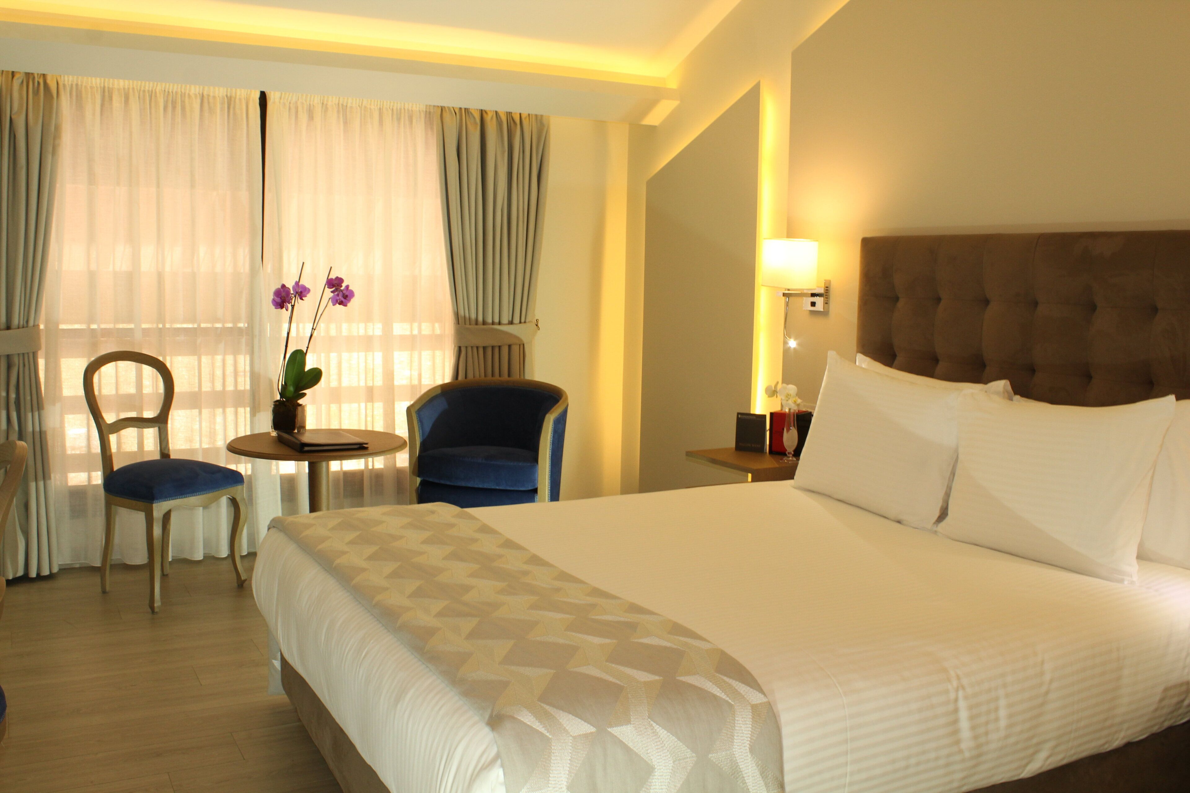 A hotel room with a large bed, a desk with a chair, a small table with a plant, and a window with curtains.