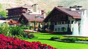 A large, multi-story building with a stone facade and a red-tiled roof, surrounded by a well-maintained garden with red flowers and a fountain. - InterContinental Mzaar Lebanon by IHG (Mzaar Kfardebian)