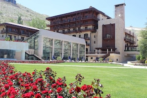 Un hotel con fachada de vidrio moderna, un jardín ajardinado con flores rojas y un telón de fondo montañoso.