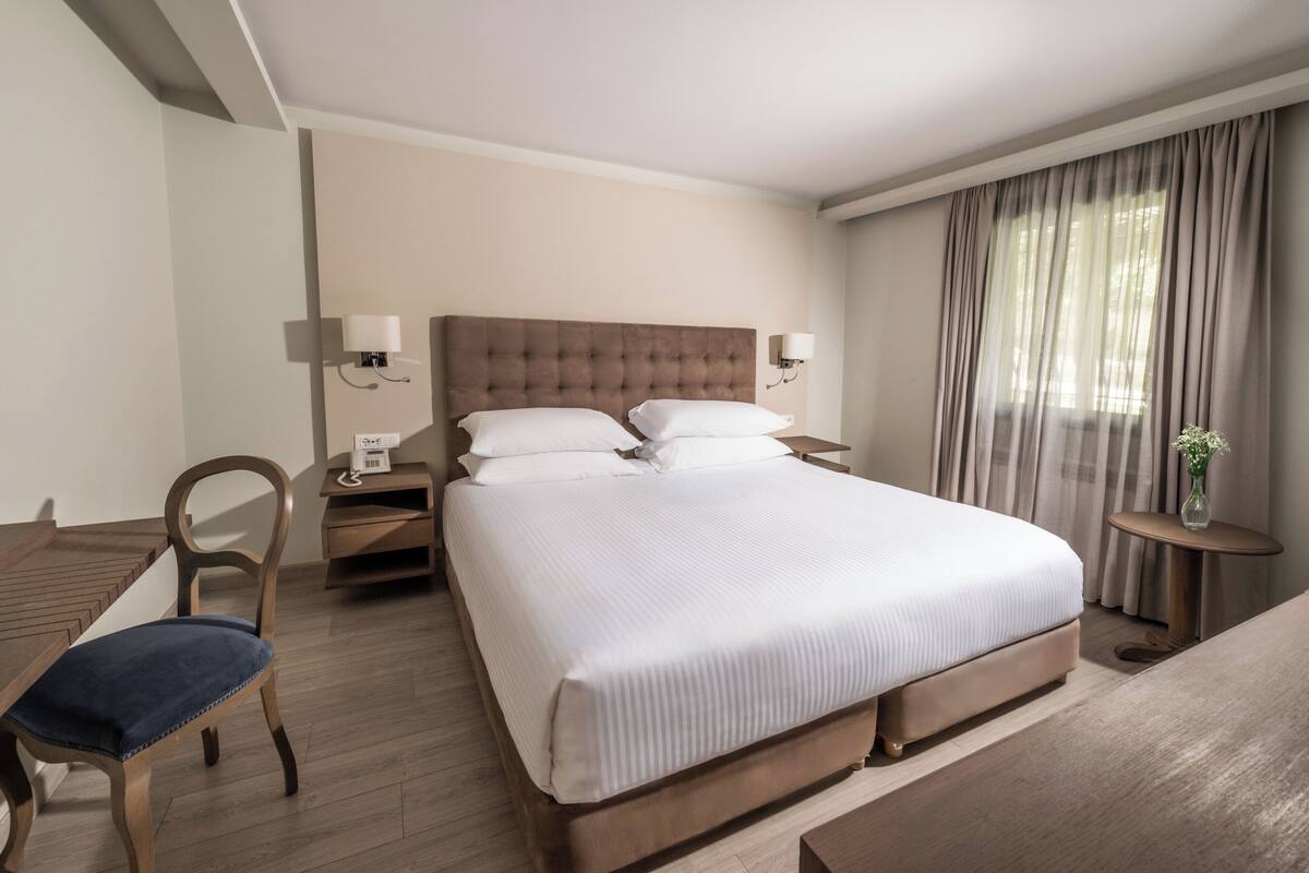 A hotel room with a large bed, a wooden desk, a chair, a bedside table, and a small table with a vase.