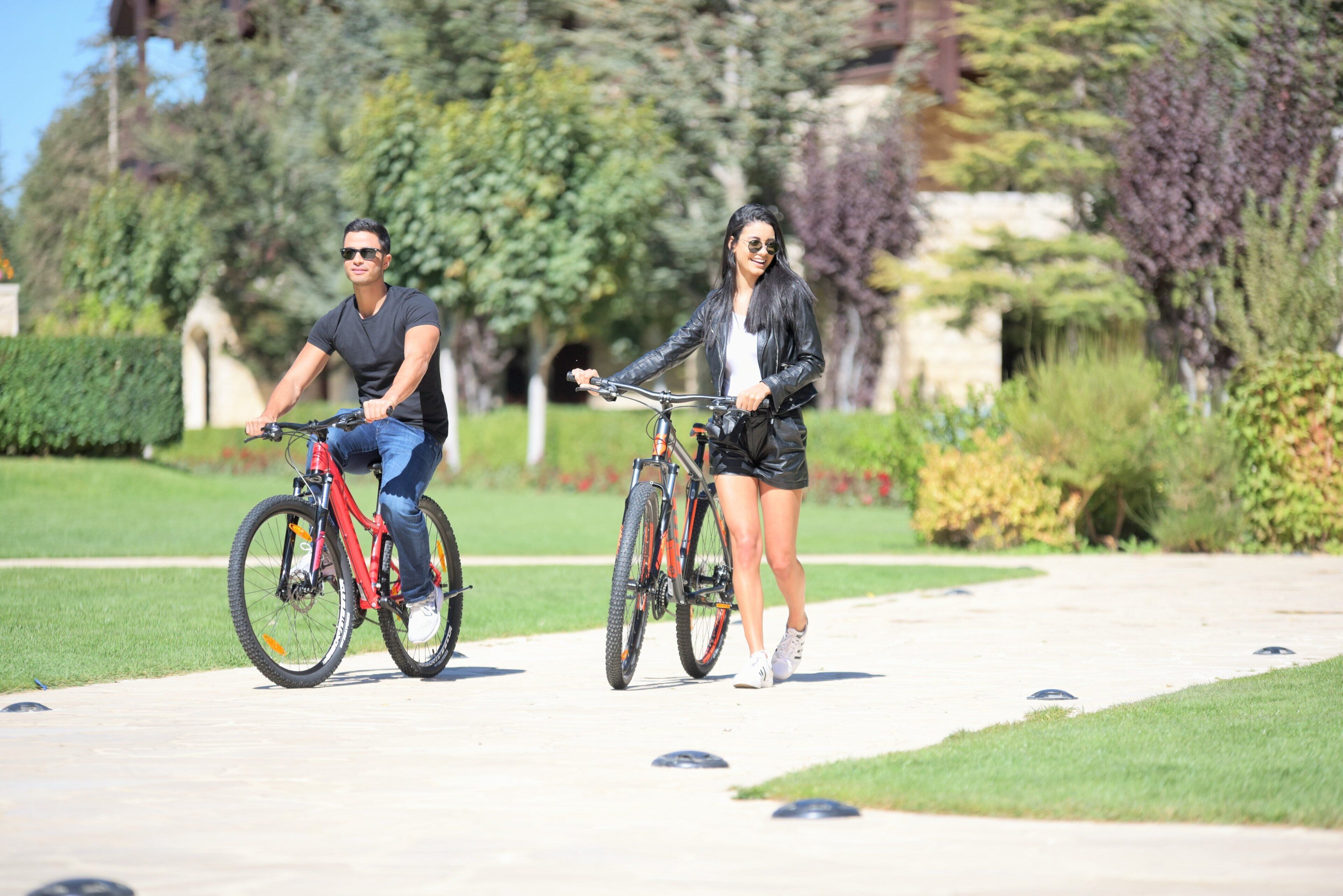 Dos personas en bicicleta en un parque con césped cuidado y árboles.