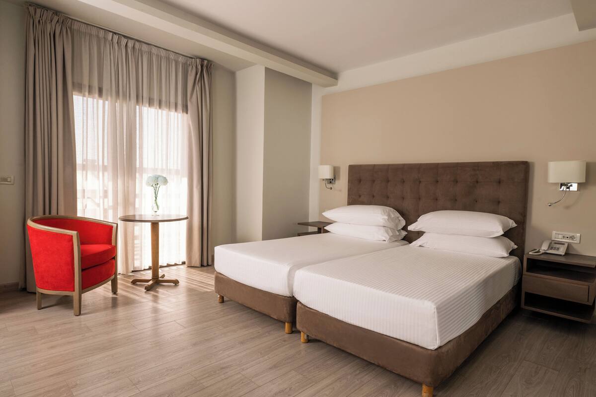 A hotel room with two beds, a red chair, a small table, and a nightstand with a phone.