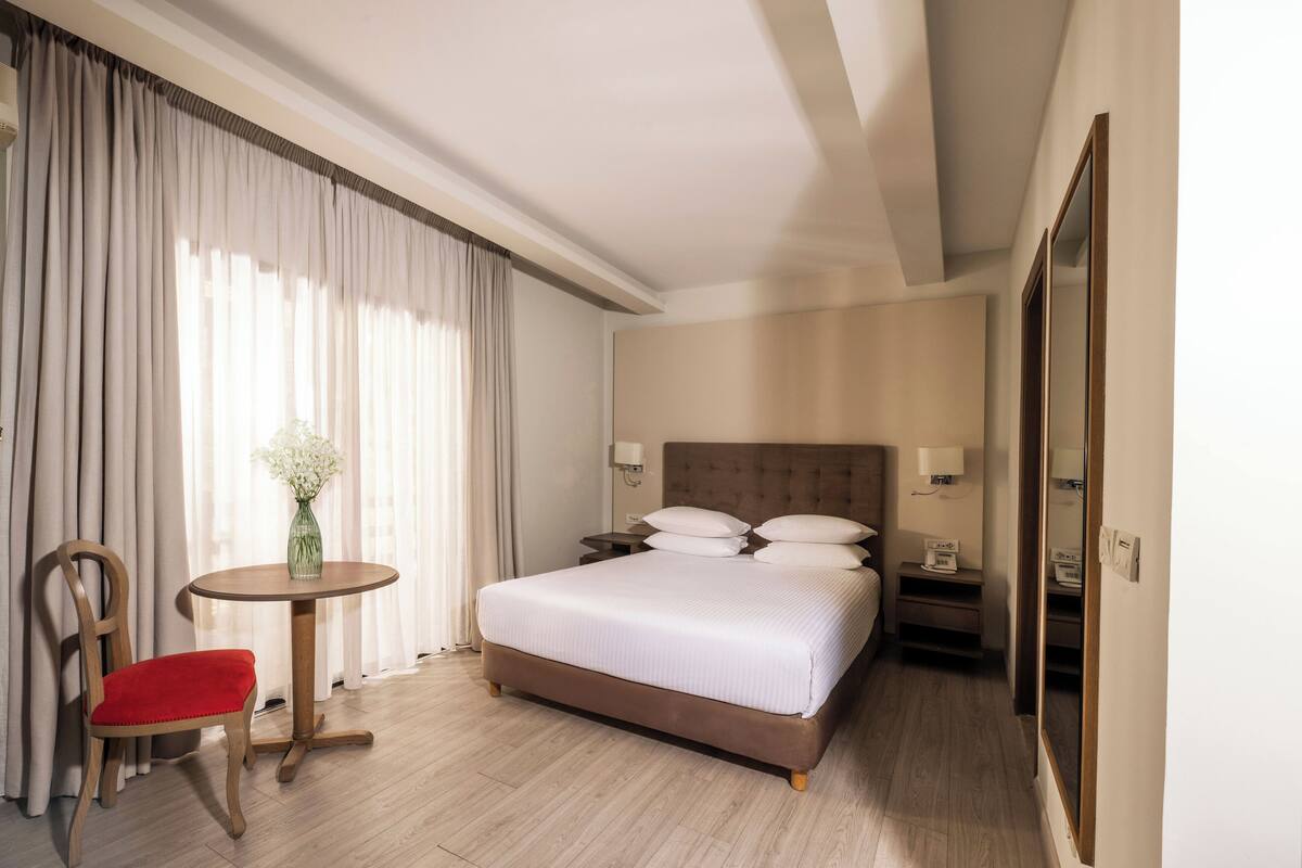A hotel room with a large bed, a wooden chair, a small round table, and a vase with flowers.