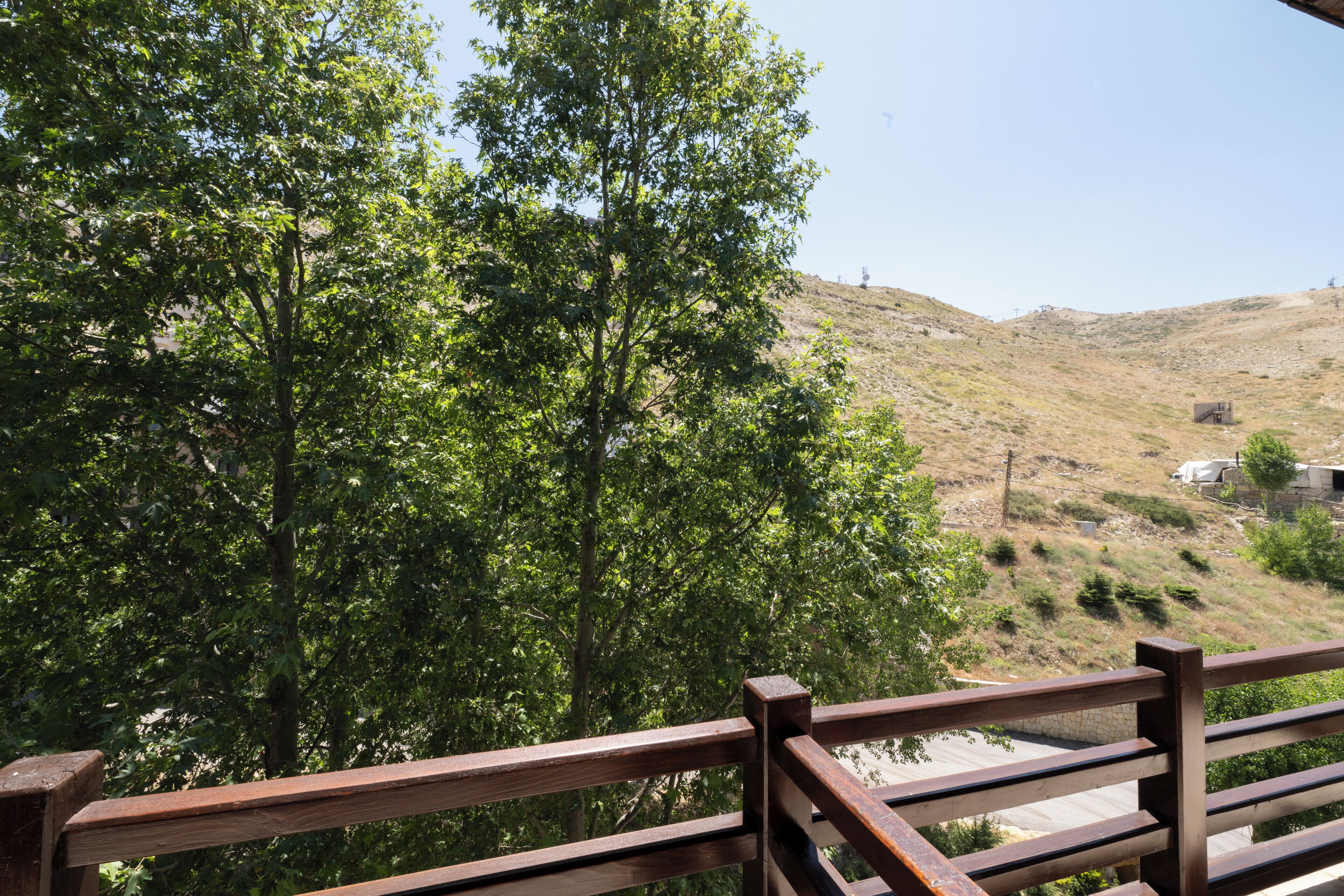 Une balustrade en bois surplombe une colline couverte d’une végétation luxuriante et verdoyante, sous un ciel dégagé.