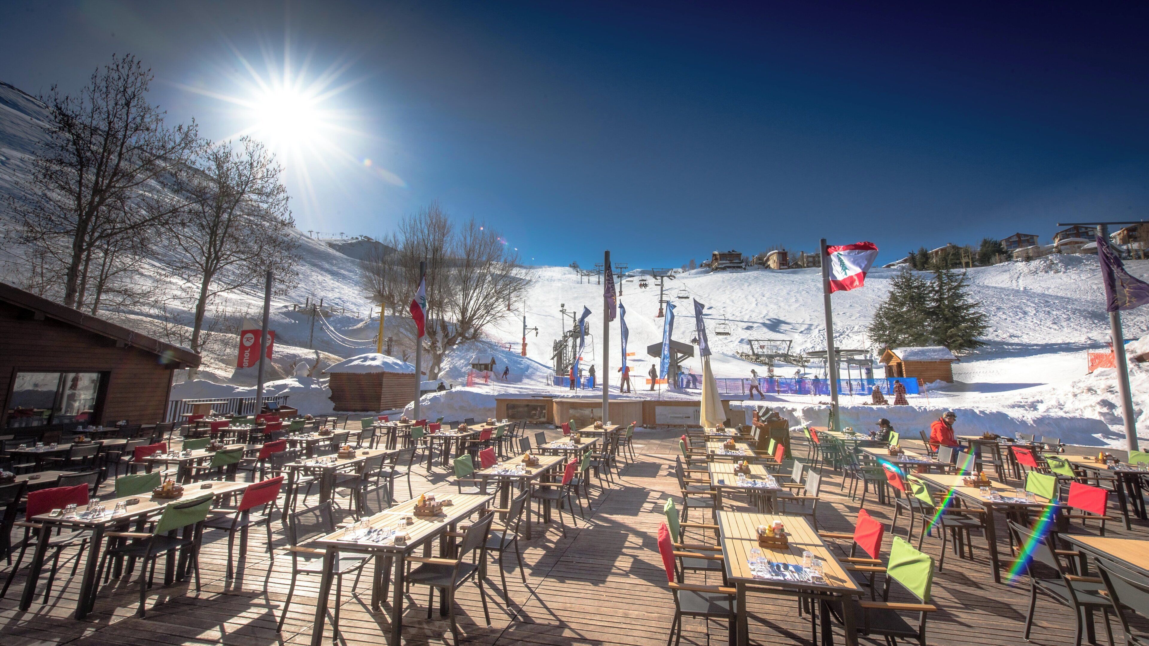 An outdoor dining area with tables and chairs, a ski slope in the background, and flags.