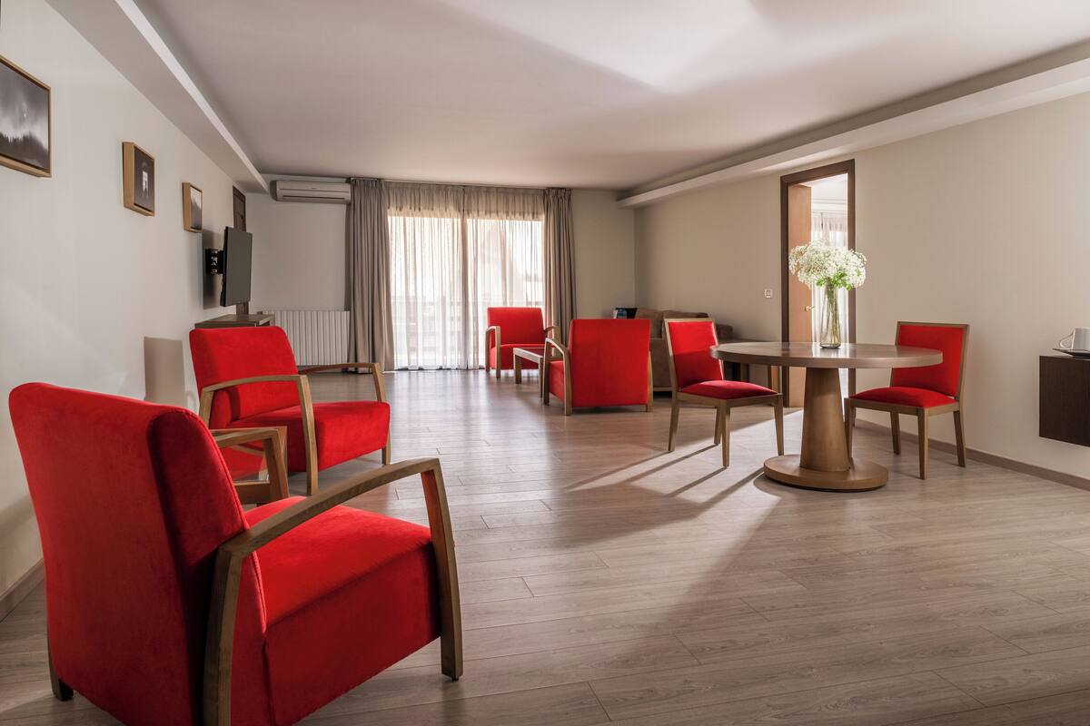 A modern hotel room with a dining area featuring red chairs and a round table, a large window with curtains, and a flat-screen TV mounted on the wall.