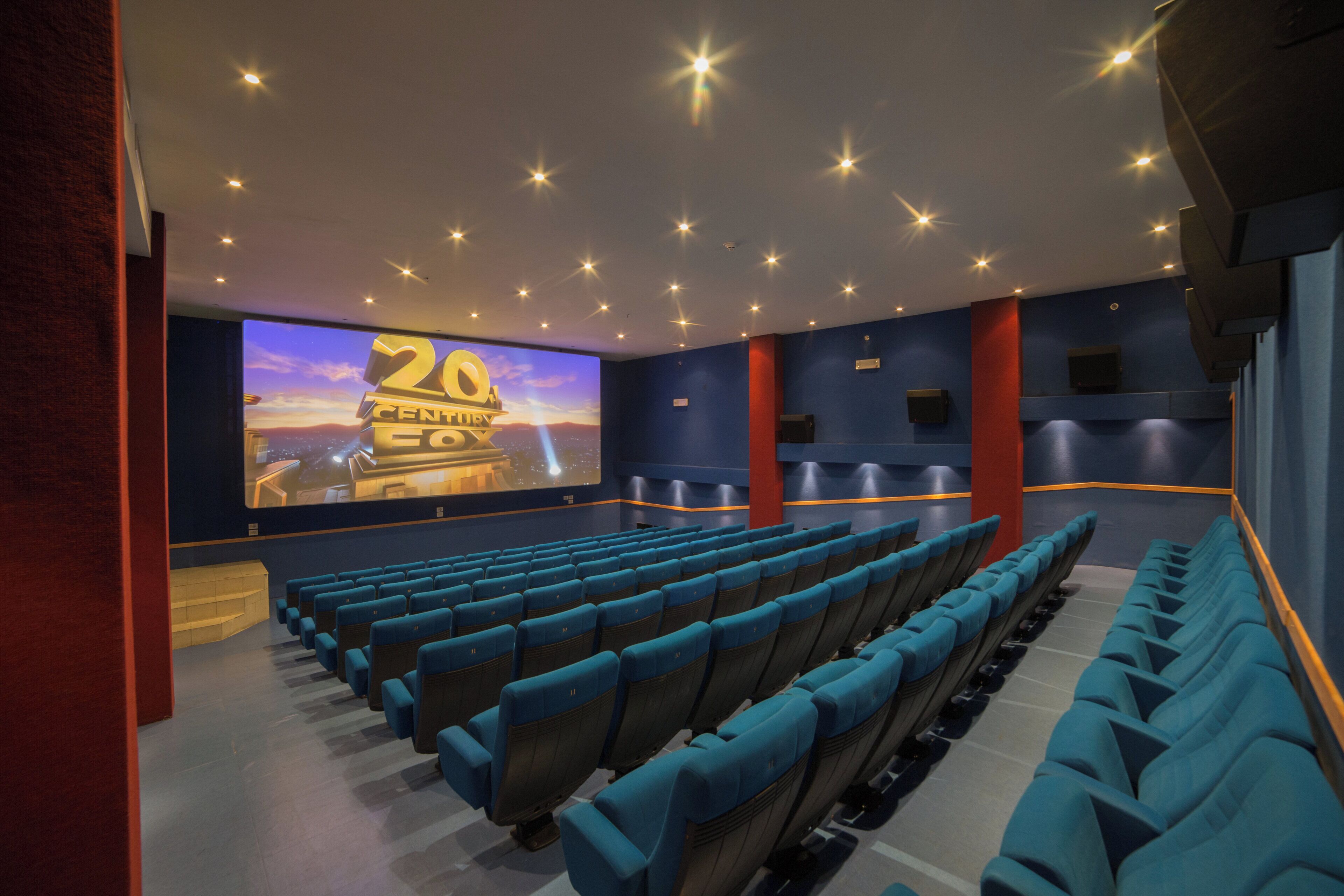 Una sala cinematografica con file di sedie blu, uno schermo grande che mostra un festival cinematografico e un moderno design del soffitto.