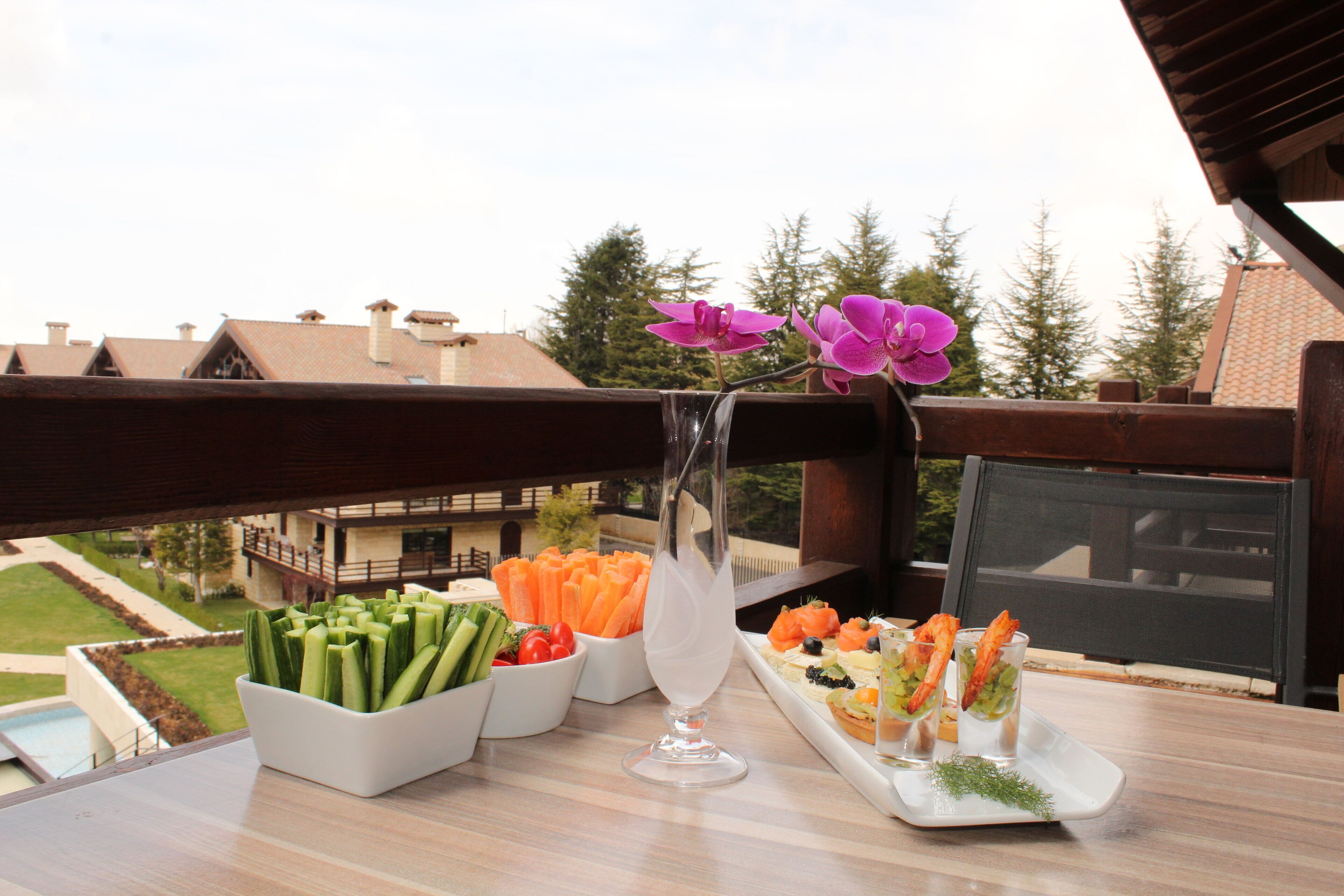 A table with food and drinks, a vase with flowers, and a view of houses and trees.