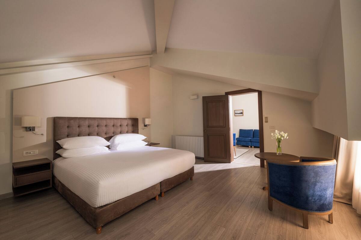 A bedroom with a large bed, a wooden floor, a blue chair, and a door leading to another room.