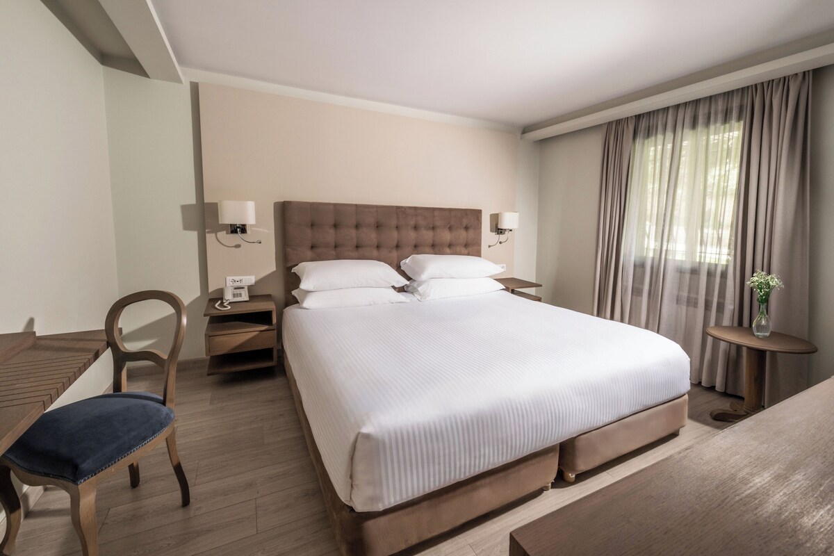 A hotel room with a large bed, a wooden desk, a chair, a bedside table, and a small table with a vase.