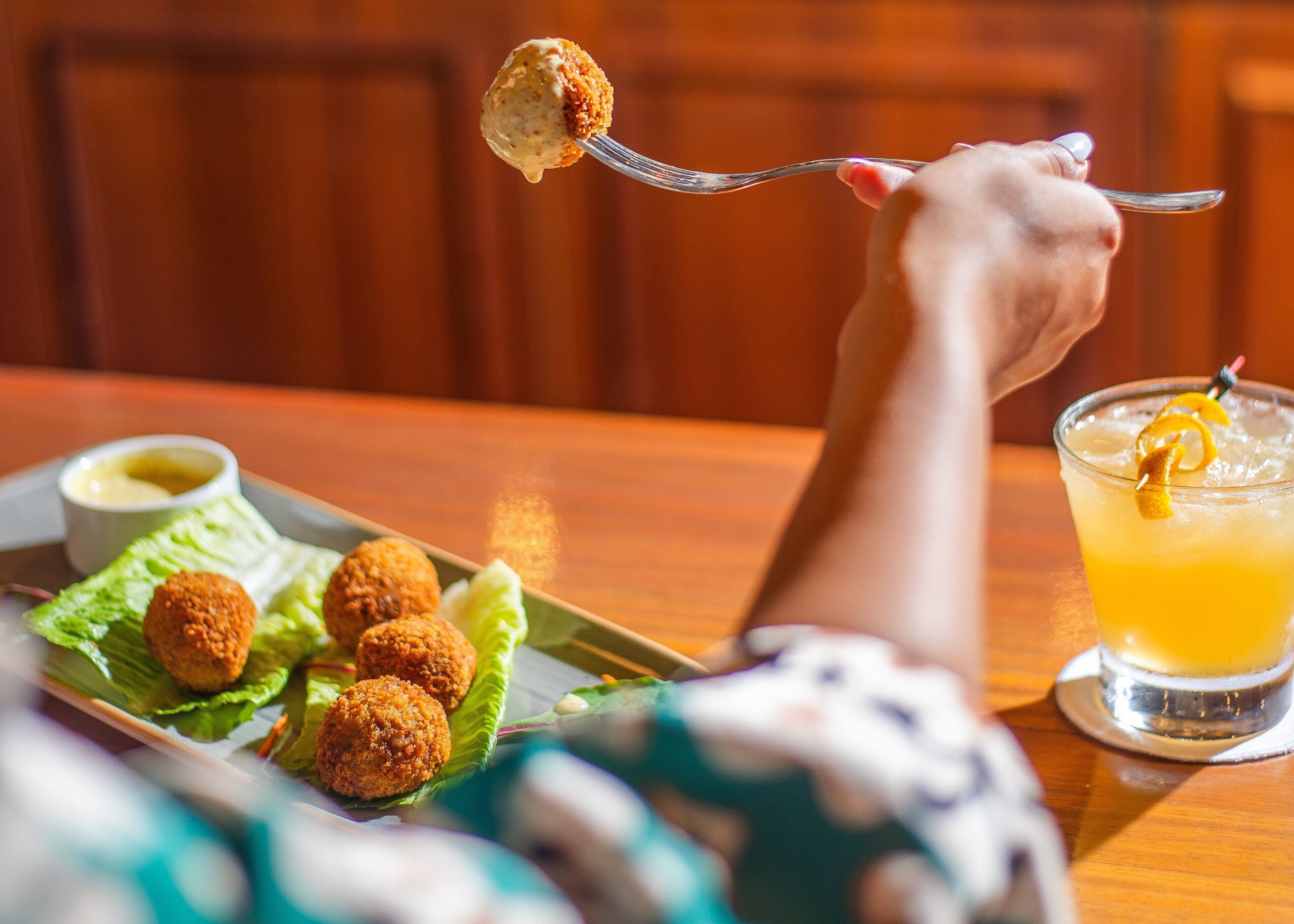 Una persona sosteniendo un tenedor con comida, un plato de comida y una bebida sobre una mesa.