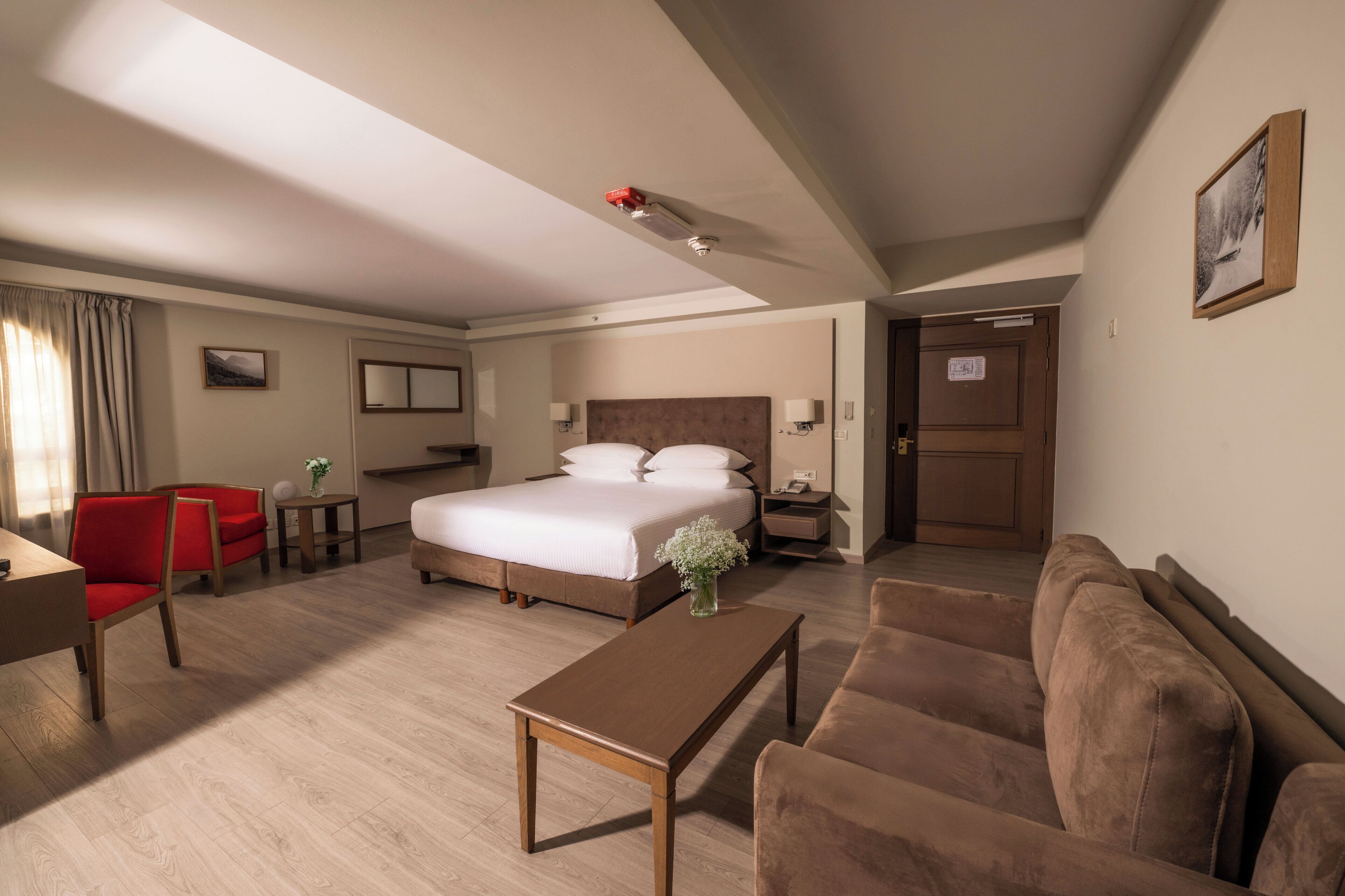 A hotel room with a bed, a red chair, a brown sofa, a wooden coffee table, and a small table with a vase.