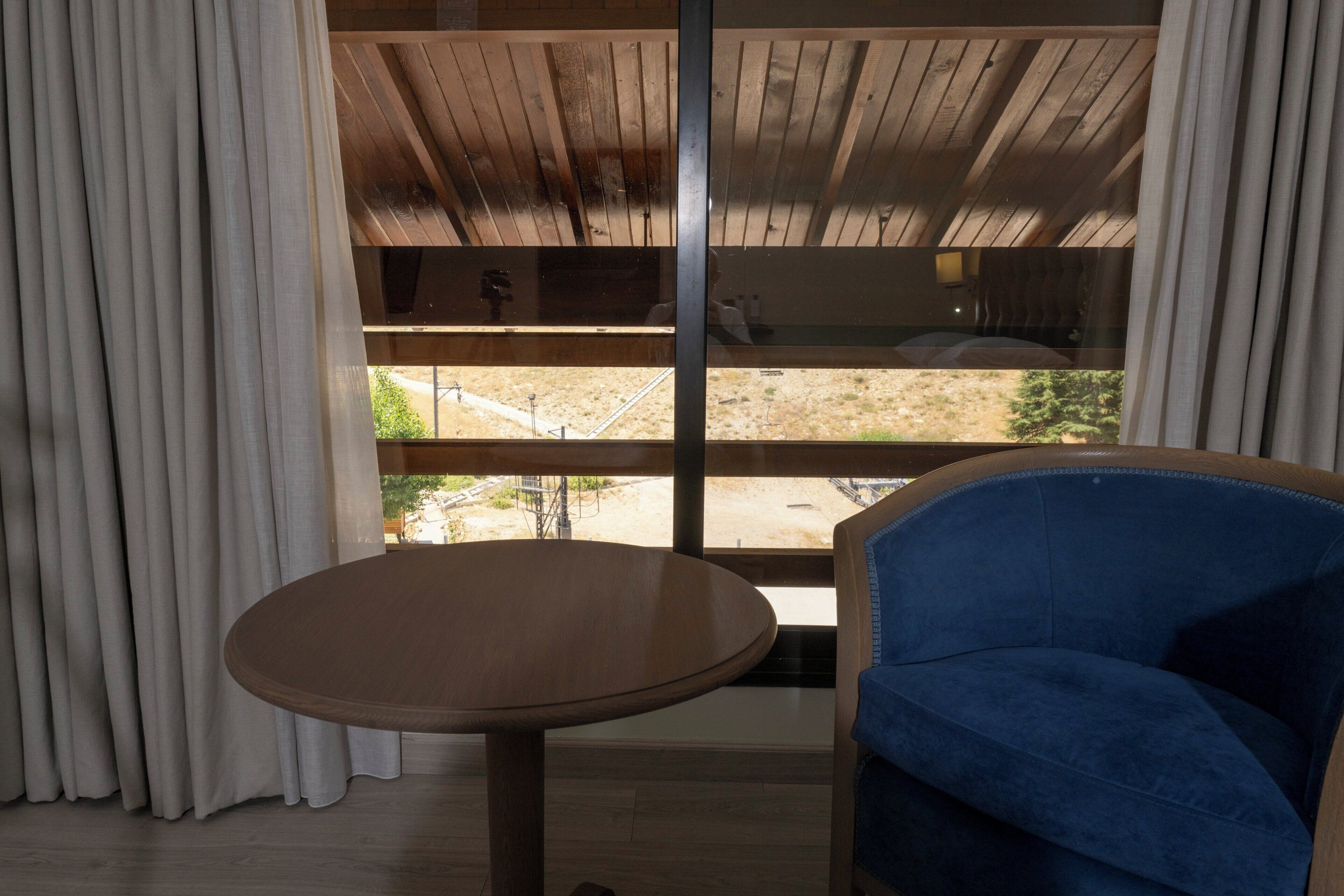 Una habitación con techo de madera, un ventanal con cortinas, una mesa redonda y una silla azul.