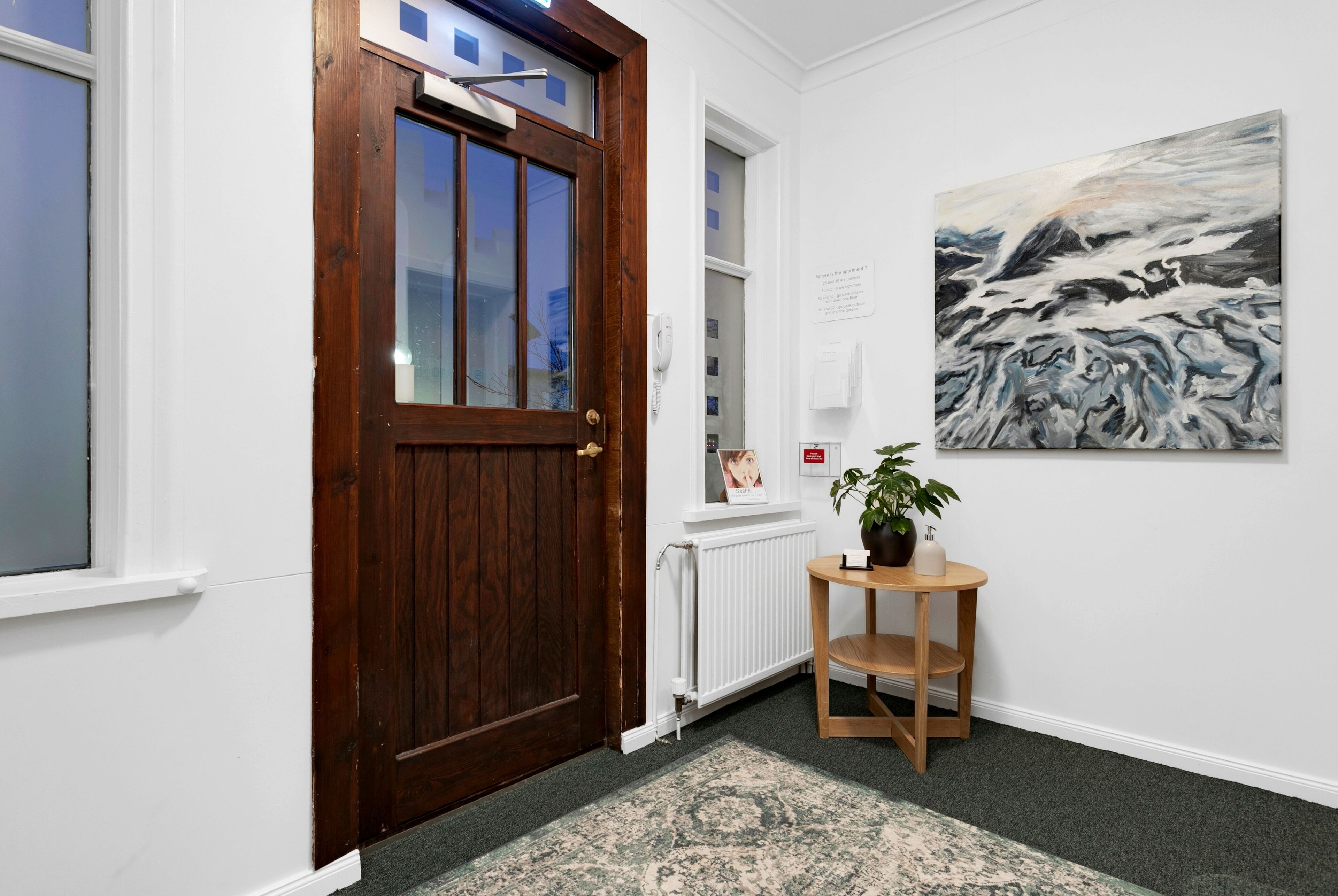Un couloir avec une porte en bois, un radiateur, une œuvre d’art encadrée et une petite table sur laquelle se trouve une plante.