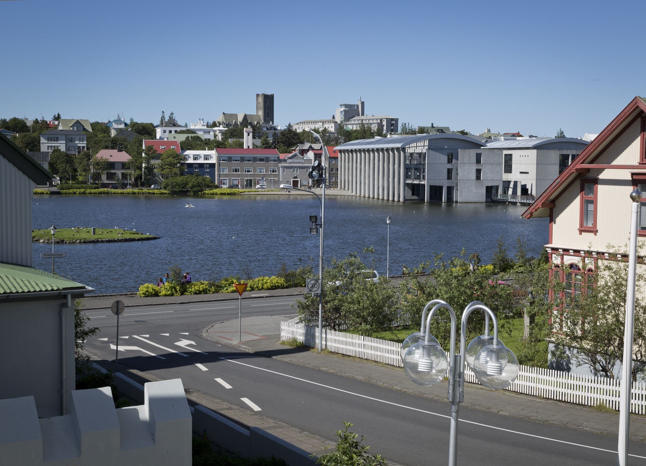 A view of a waterfront street with buildings, a body of water, and a white fence.