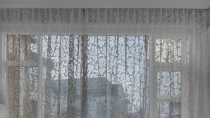 Ett rum med två grå fåtöljer med vita kuddar, ett litet runt bord med ett glas och en grön kopp, och ett fönster med tunna, genomskinliga gardiner.
