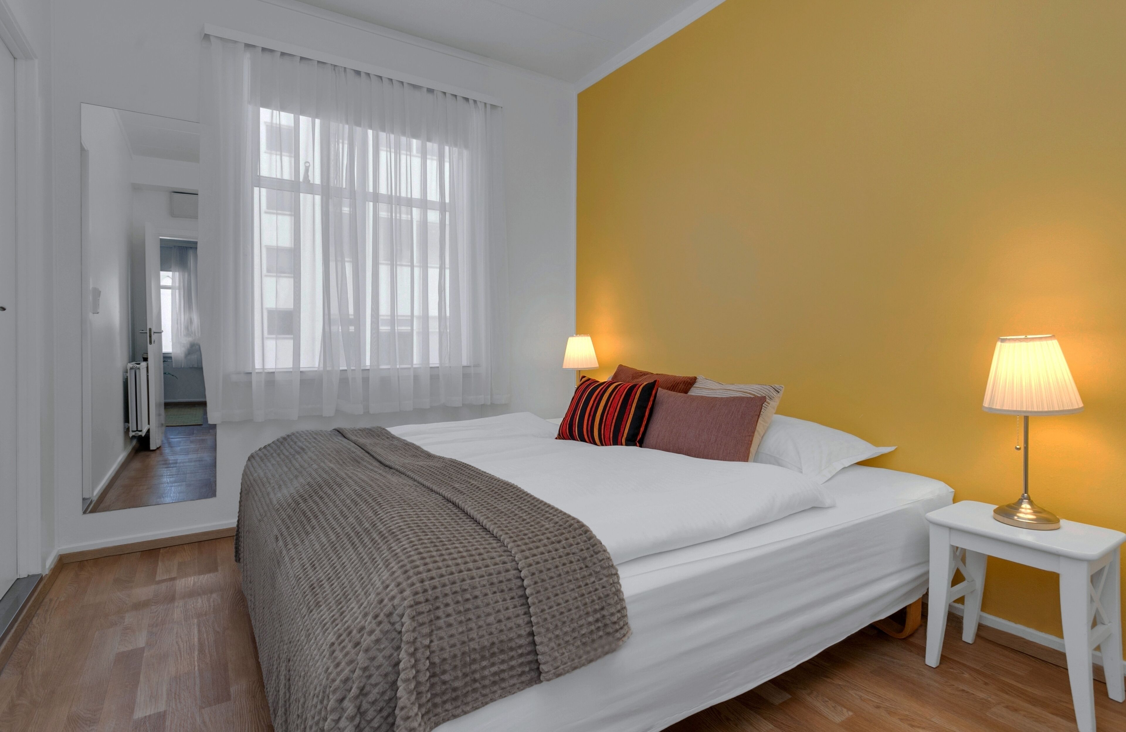 A hotel room with a large bed, a bedside table with a lamp, a window with sheer curtains, and a view of the hallway.