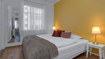 A hotel room with a large bed, a bedside table with a lamp, a window with sheer curtains, and a view of the hallway.