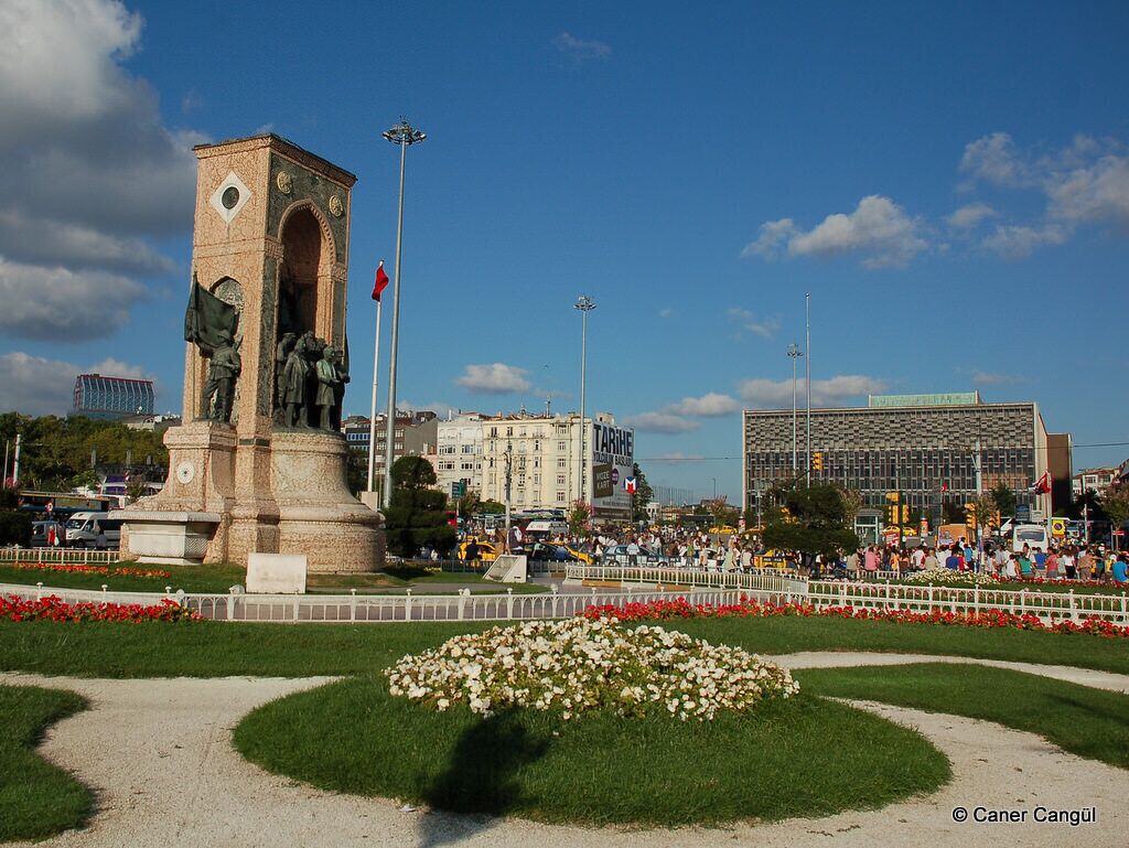 Un monumento con una estatua, rodeado de un macizo de flores y una multitud de personas.