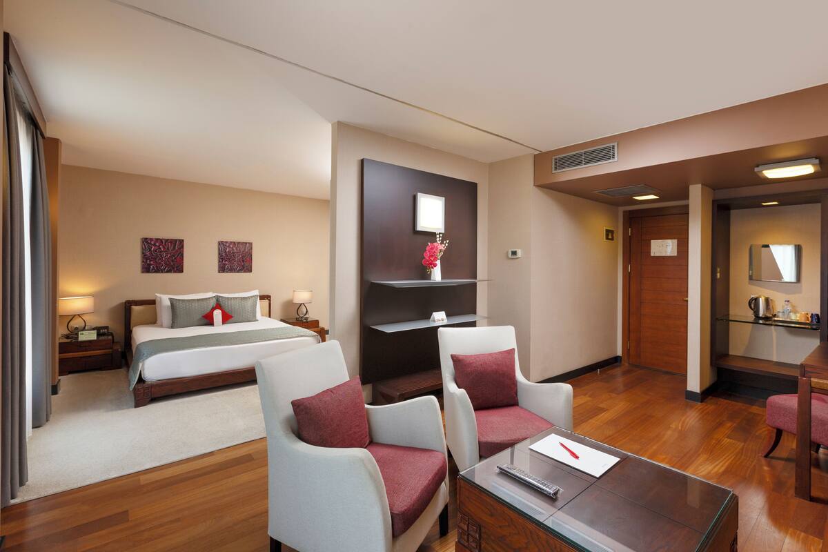 A hotel room with a bed, two armchairs, a desk with a glass top, a small table, a nightstand, a lamp, a wall-mounted shelf with a vase, and a door leading to another room.