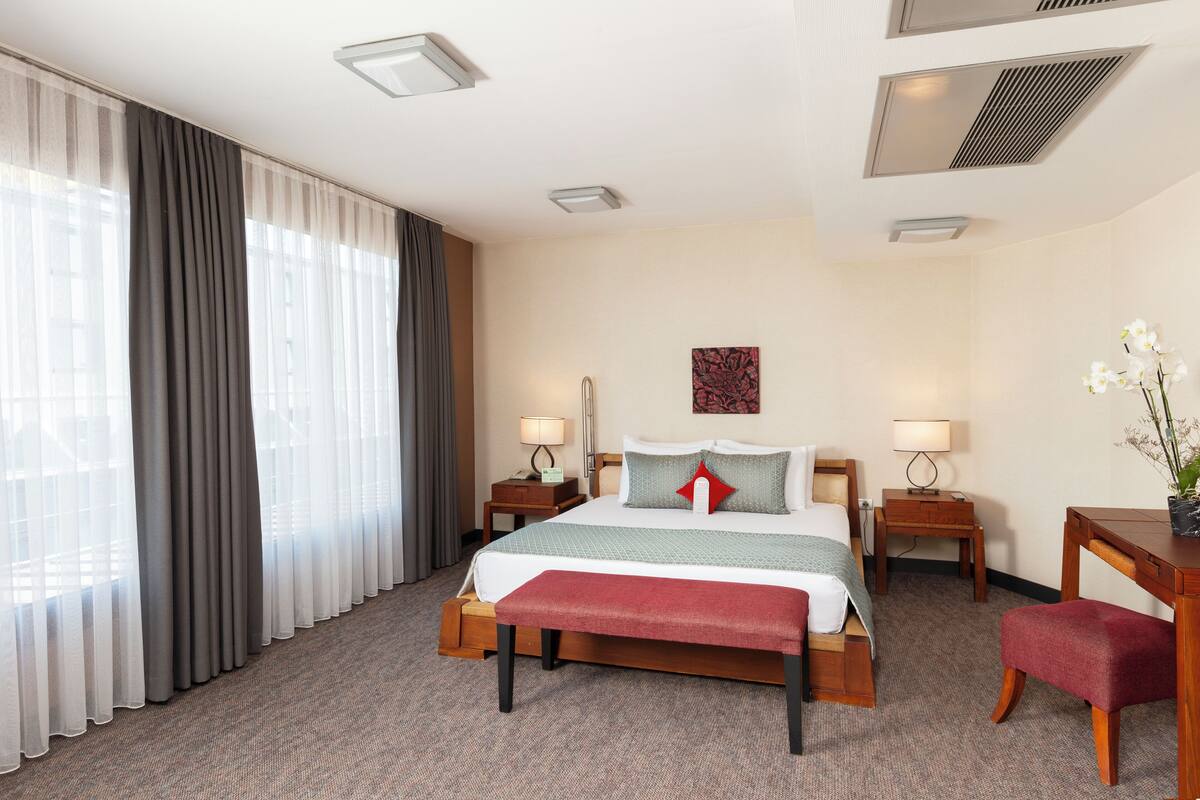 A hotel room with a large bed, two bedside tables with lamps, a desk with a chair, and a large window with curtains.