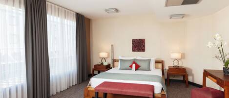 A hotel room with a large bed, two bedside tables with lamps, a desk with a chair, and a large window with curtains.