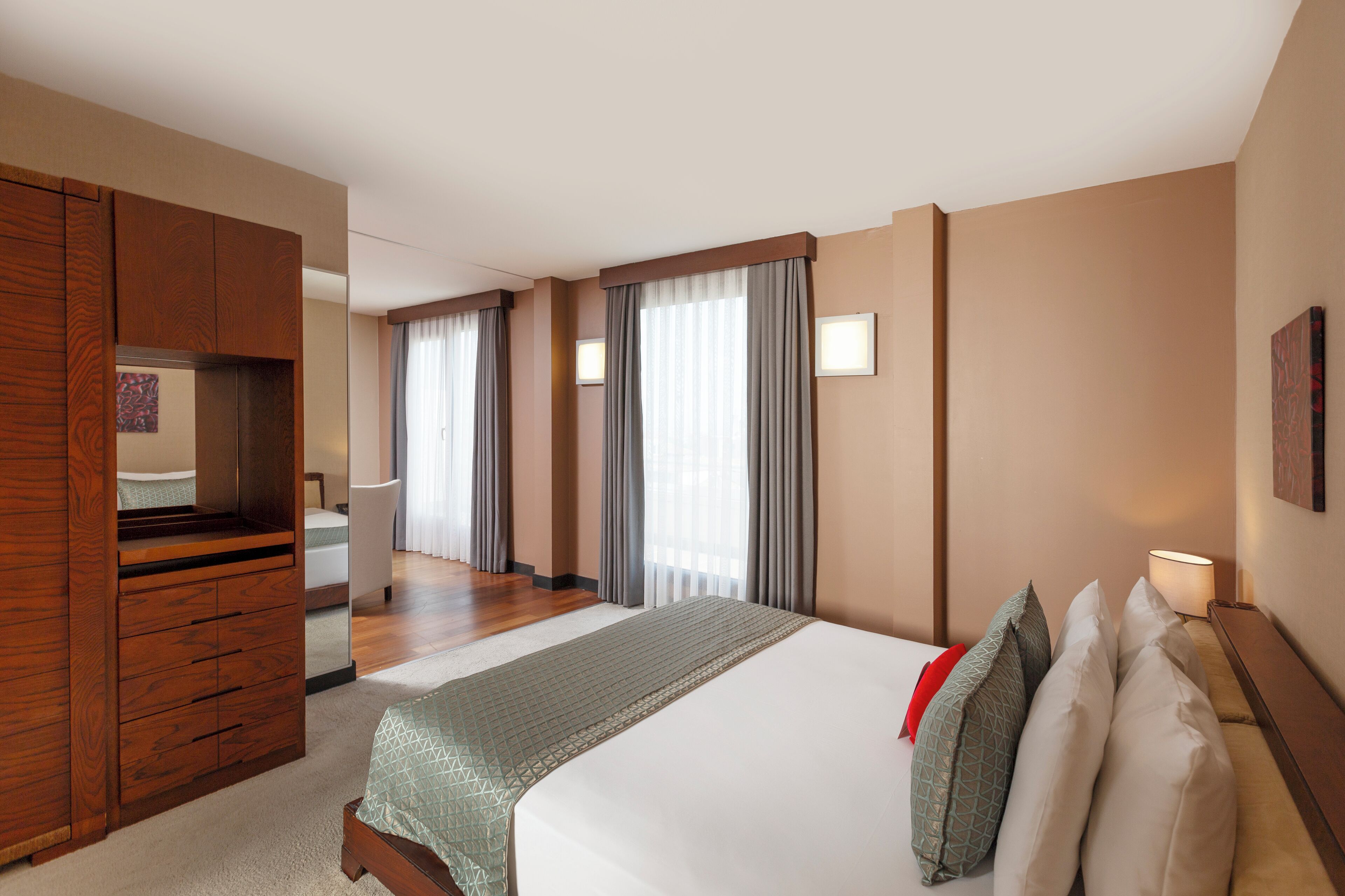 Habitación de hotel con una cama grande, un armario de madera, un escritorio con una silla y un ventanal con cortinas.