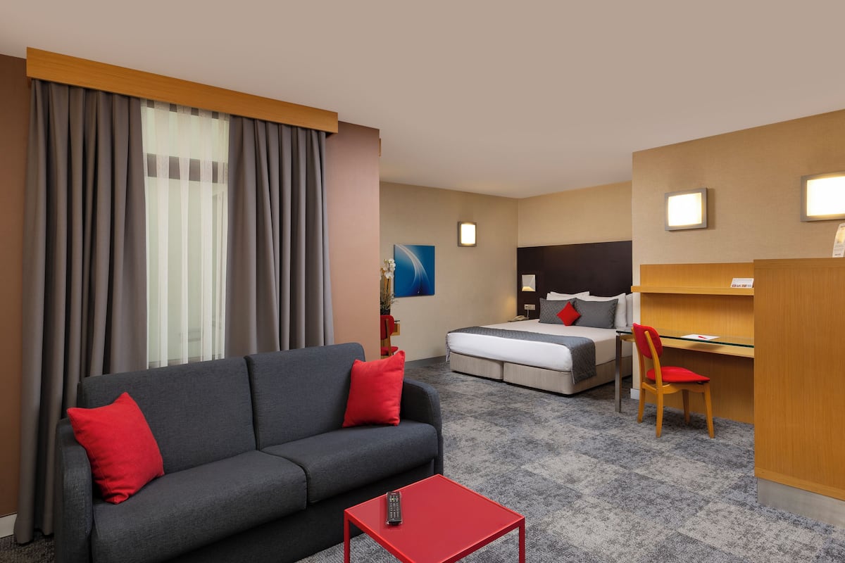 A hotel room with a gray sofa, red pillows, a red coffee table, a bed, a desk, and a chair.